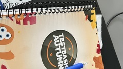 A notebook with the test bash autumn logo placed on a laptop. Also visible a phone, pen and sticker for Leading testing activities  image