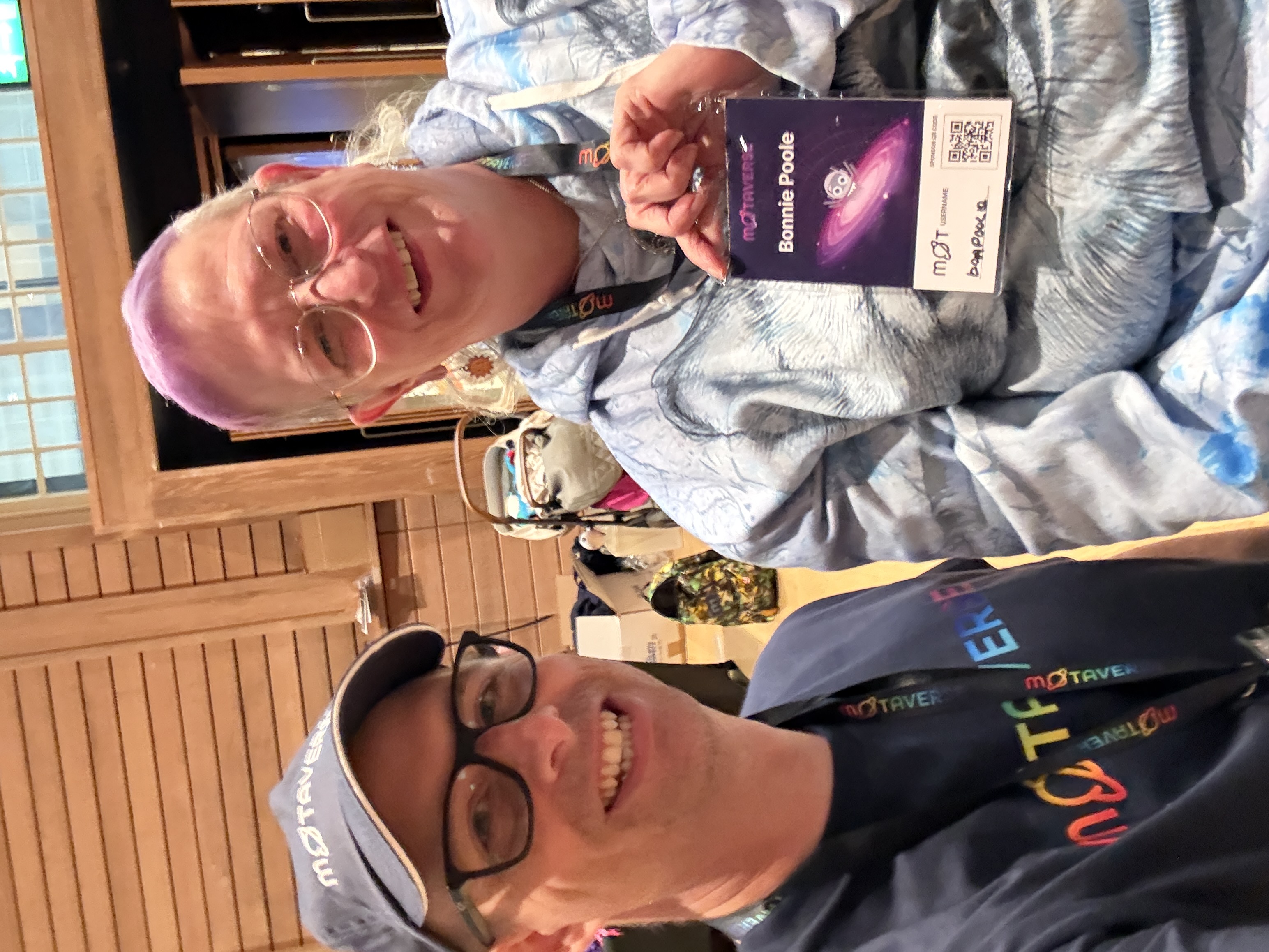 A man wearing a dark blue MoTaverse shirt and a cap is taking a selfie with a smiling woman. The woman has light purple hair, wears glasses, and a light blue and white tie-dye-patterned hoodie. She is holding up a conference badge with her name, Bonnie Poole, visible against a space-themed background. They are indoors in a room with light wooden paneling.