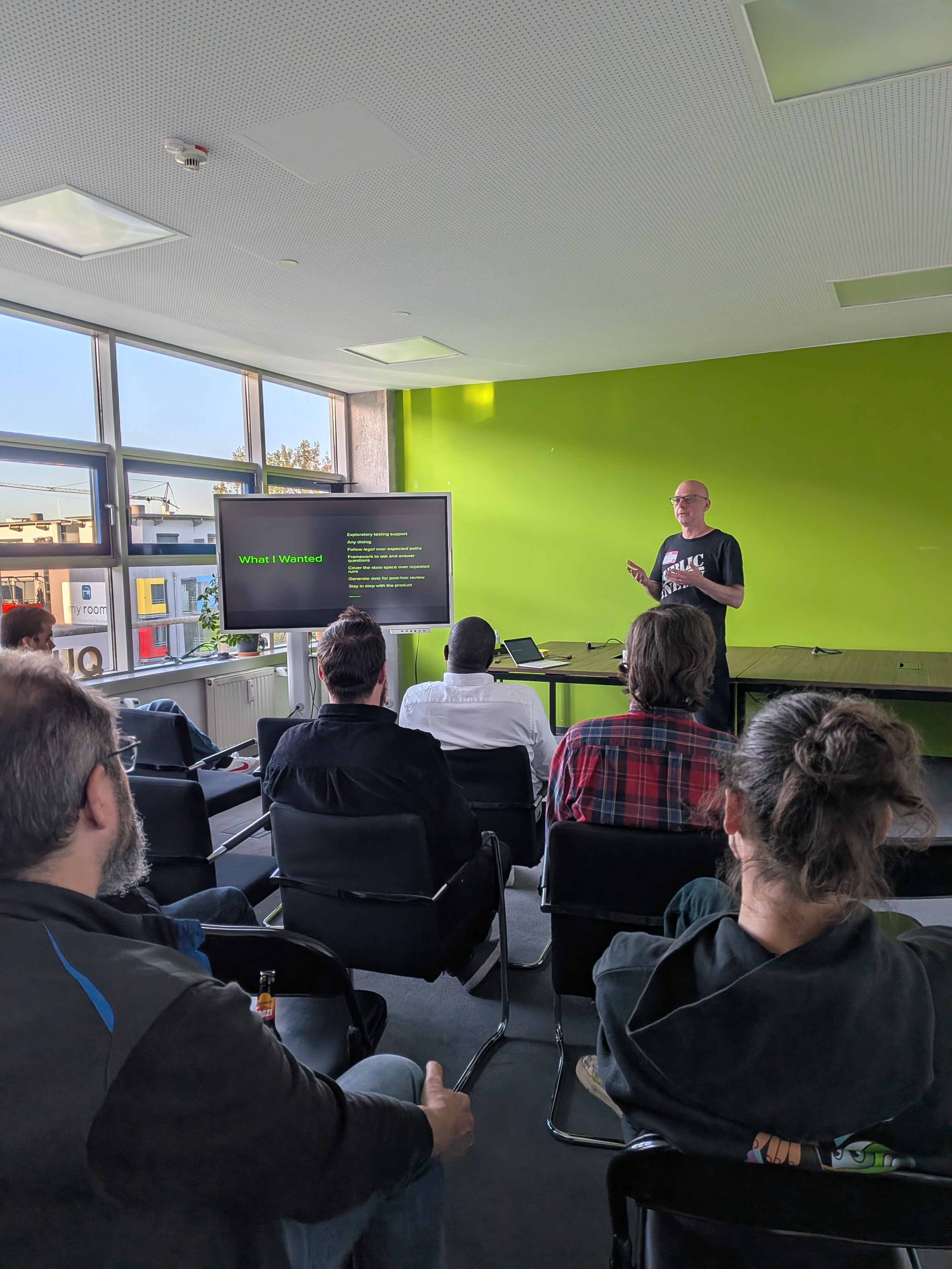 A person gives a presentation to a seated audience in a room with a green wall and large windows, displaying slides o... image