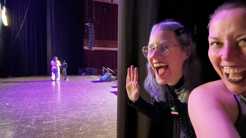 Veerle and Gwen laugh and wave in a backstage selfie while Susanne speaks at a lectern on the purple-lit stage behind... image