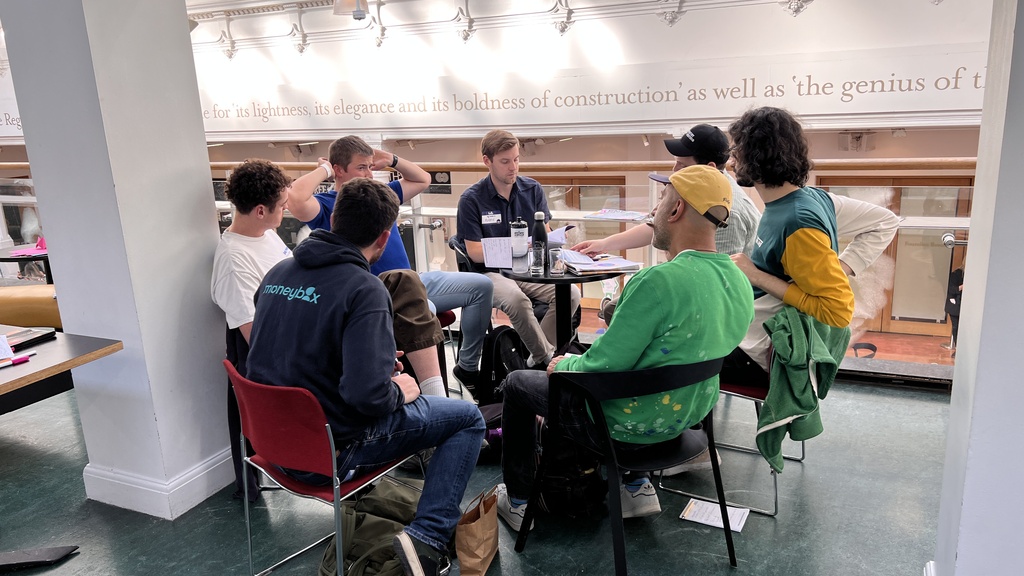 A group of people sitting in a circle of chairs having a collaborative group discussion around small tables. In the f... image