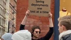 A person in a black shirt holds up a sign against a red brick wall in an urban setting. The sign reads 'I'm QA Superhero: Saving deployments one test at a time'. Several people wearing winter hats ... image