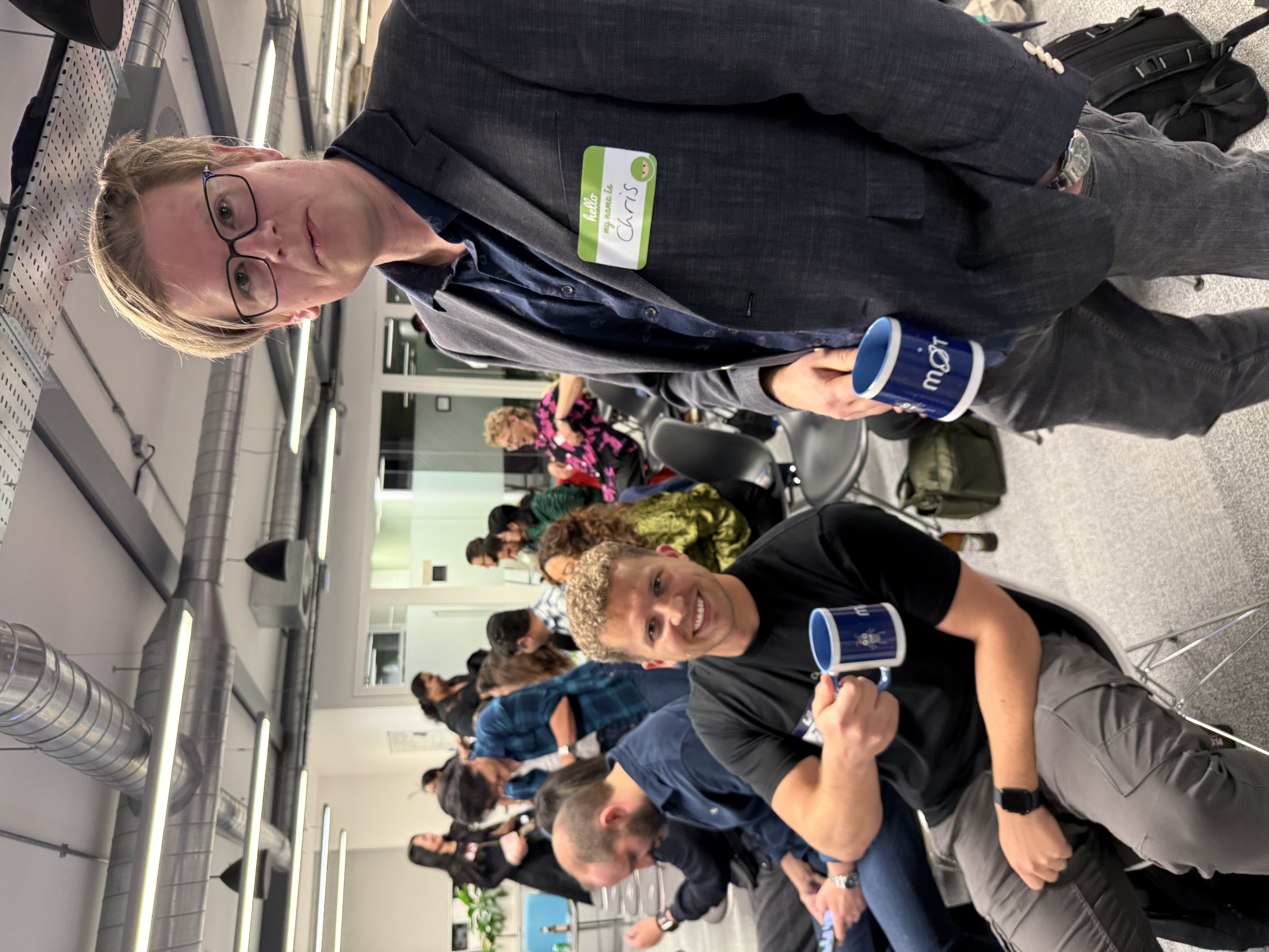 Two men are in the foreground, holding mugs and looking towards the camera. The man on the right is wearing glasses, a grey blazer, a dark blue shirt, and grey trousers. He has a name tag that reads "Hello, My name is Chris". The man on the left is smiling and wearing a black t-shirt, grey trousers, and a black smartwatch. He is sitting on a black chair. Both mugs are white with a blue rim and blue text. There is a bug character on the mug and an MoT logo. 

In the background, several other people are visible, some sitting and some standing, engaged in conversation. The setting appears to be an informal gathering or a casual office environment with exposed ceiling ducts and fluorescent lighting. There are also chairs and bags scattered in the background.