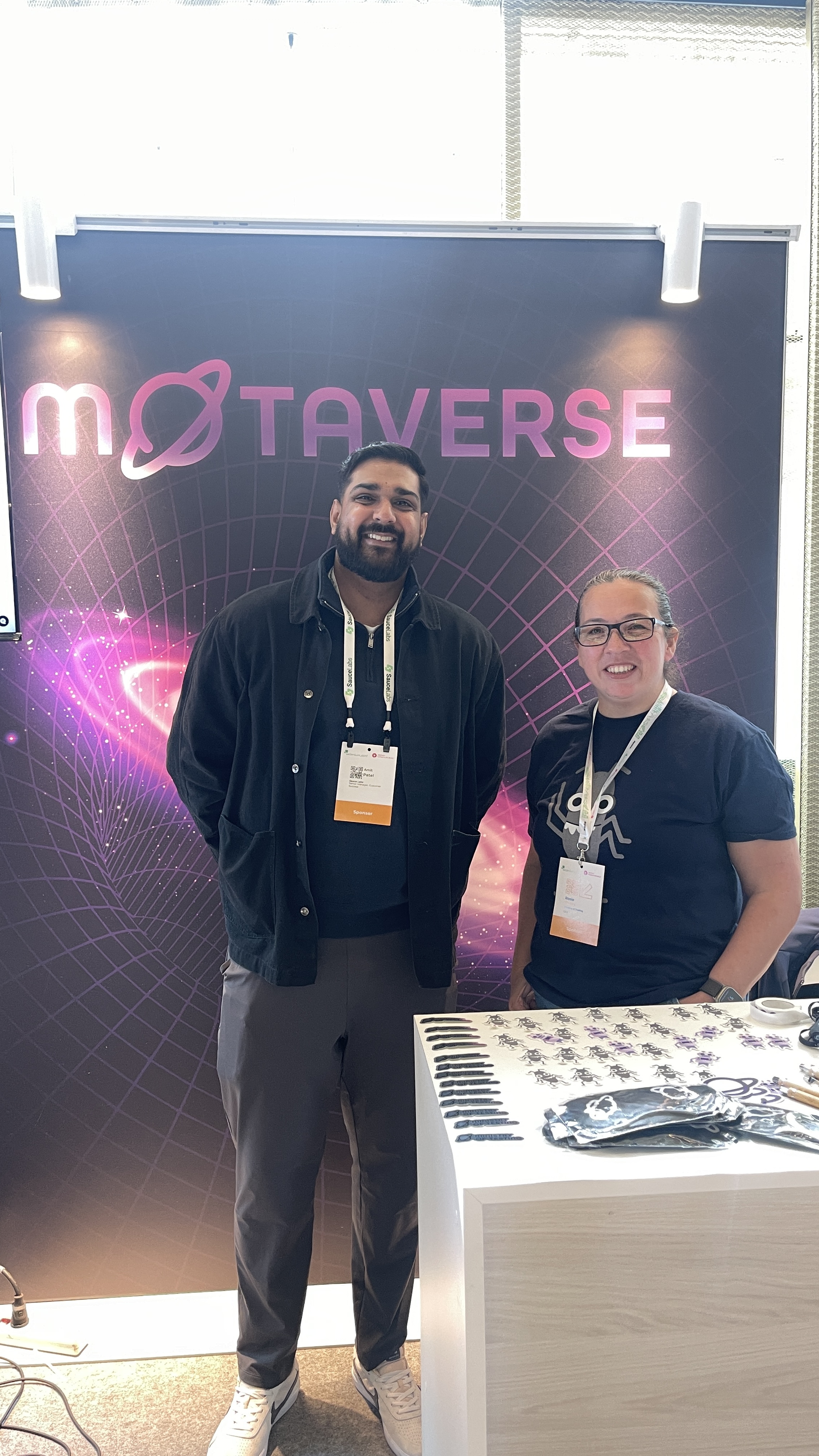Amit Patel from Sauce Labs stands smiling next to Rosie Sherry at the MoTaverse stand at Selenium Conference. The stand is decorated with bug-themed stickers and Ministry of Testing branding.