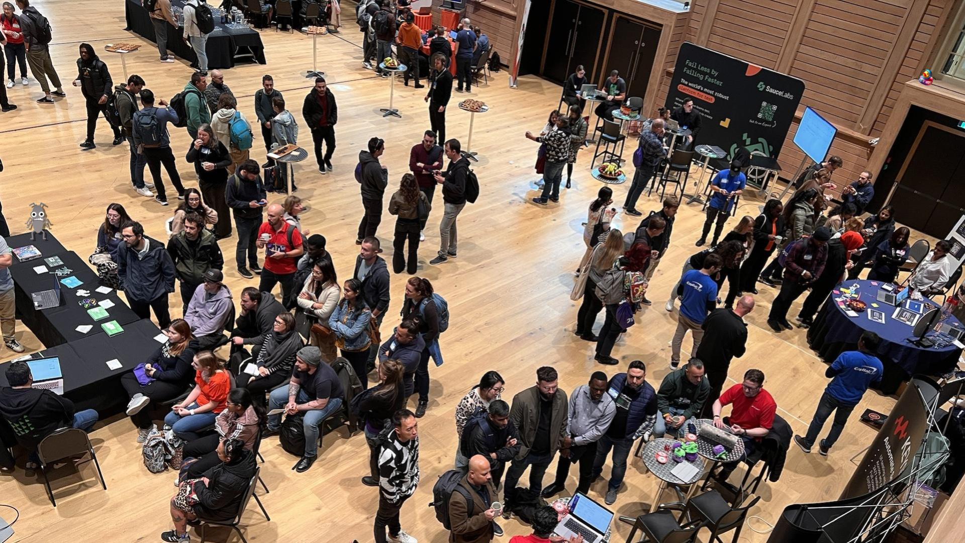 Looking down on crowds of people gathered around displays from companies in the testing community. There appears to be lots going on. There is a small bug character and duck character immersed in the scene.