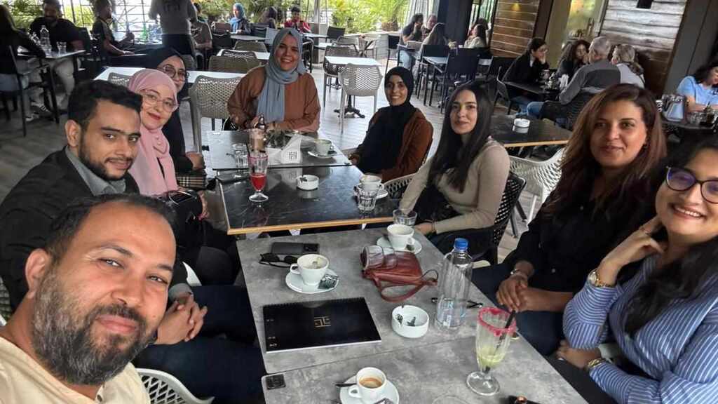 A group of nine people, including both men and women, are gathered around a table at an outdoor café. They are smiling and appear to be enjoying a casual meet-up or discussion. On the table are coffee cups, glasses, notebooks, a water bottle, and a purse. The background shows other patrons seated at tables, lush greenery, and a shaded terrace area with natural light filtering in.