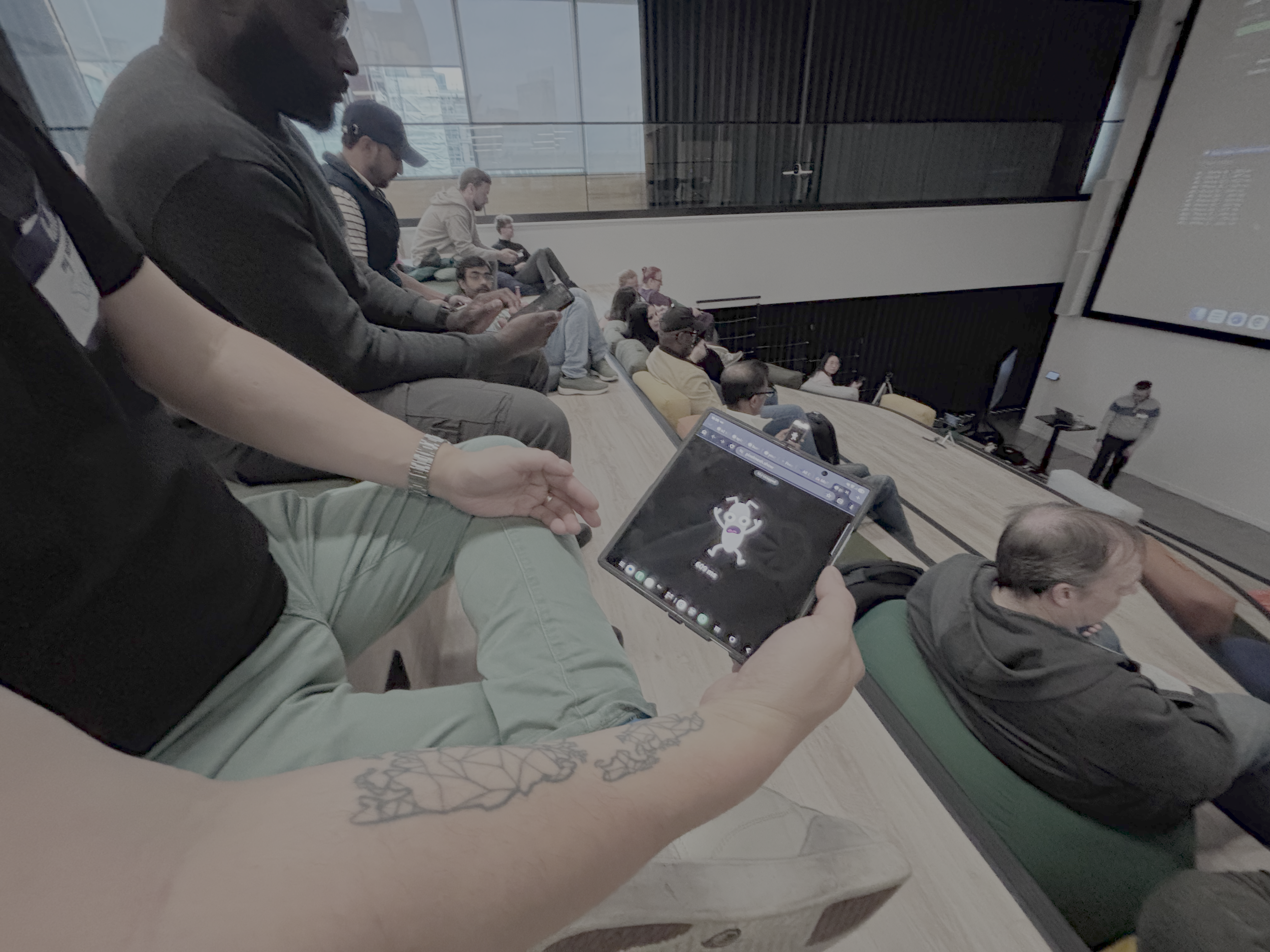 A person with an arm tattoo sits in a tiered auditorium-style seating area, holding a foldable tablet that displays a... image
