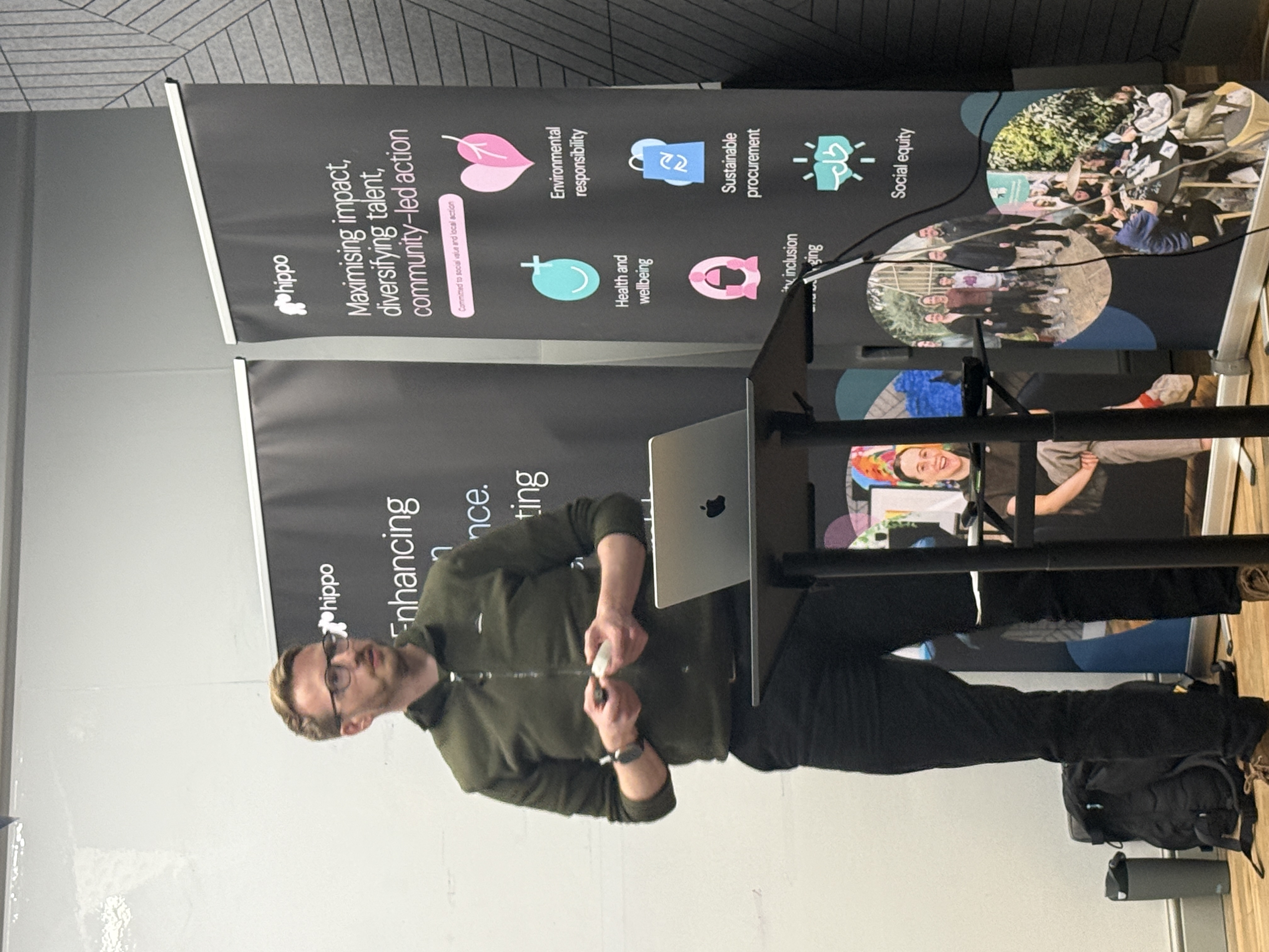 Adam who is a tall white male in black trousers and an olive green shirt is stool at the front delivering his talk.  image