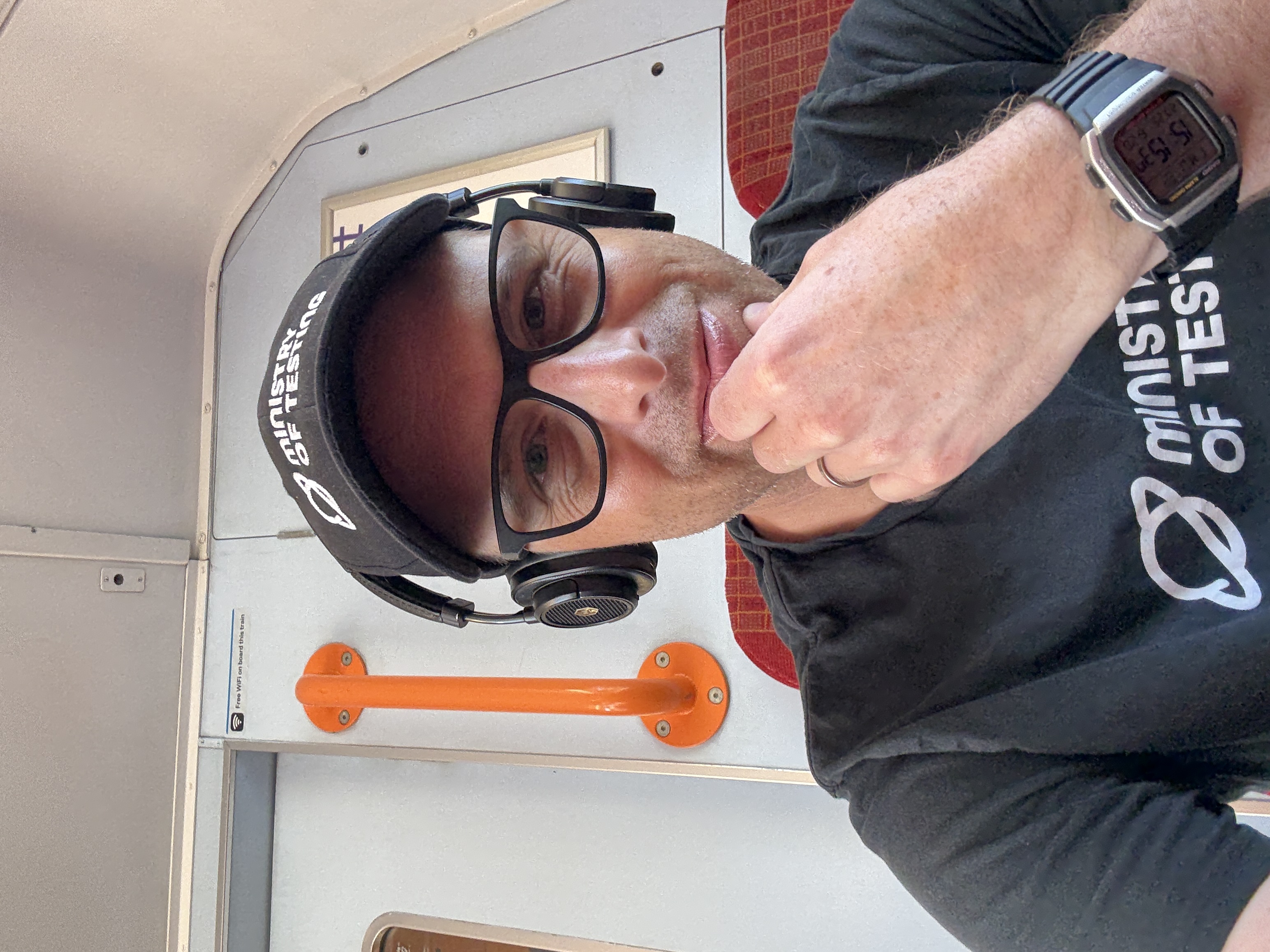 A selfie of a middle aged man with a black t-shirt and cap sat on a train. 