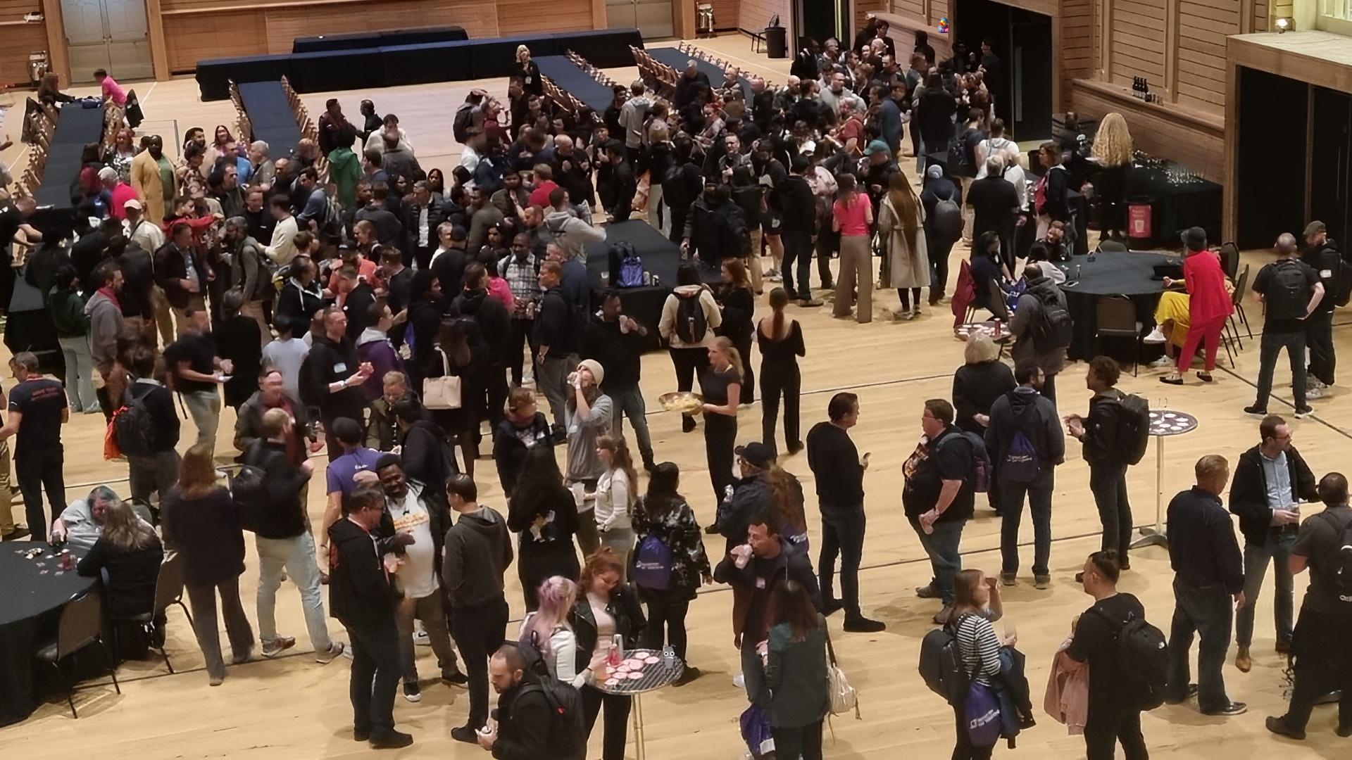 A large crowd of people gathered in a grand event space. Lots of conversations are happening. A duck and bug character are hiding.
