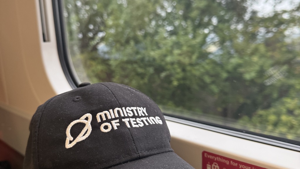 A ministry of testing branded black baseball cap sits on a lunch box next to the train window with the countryside pa... image