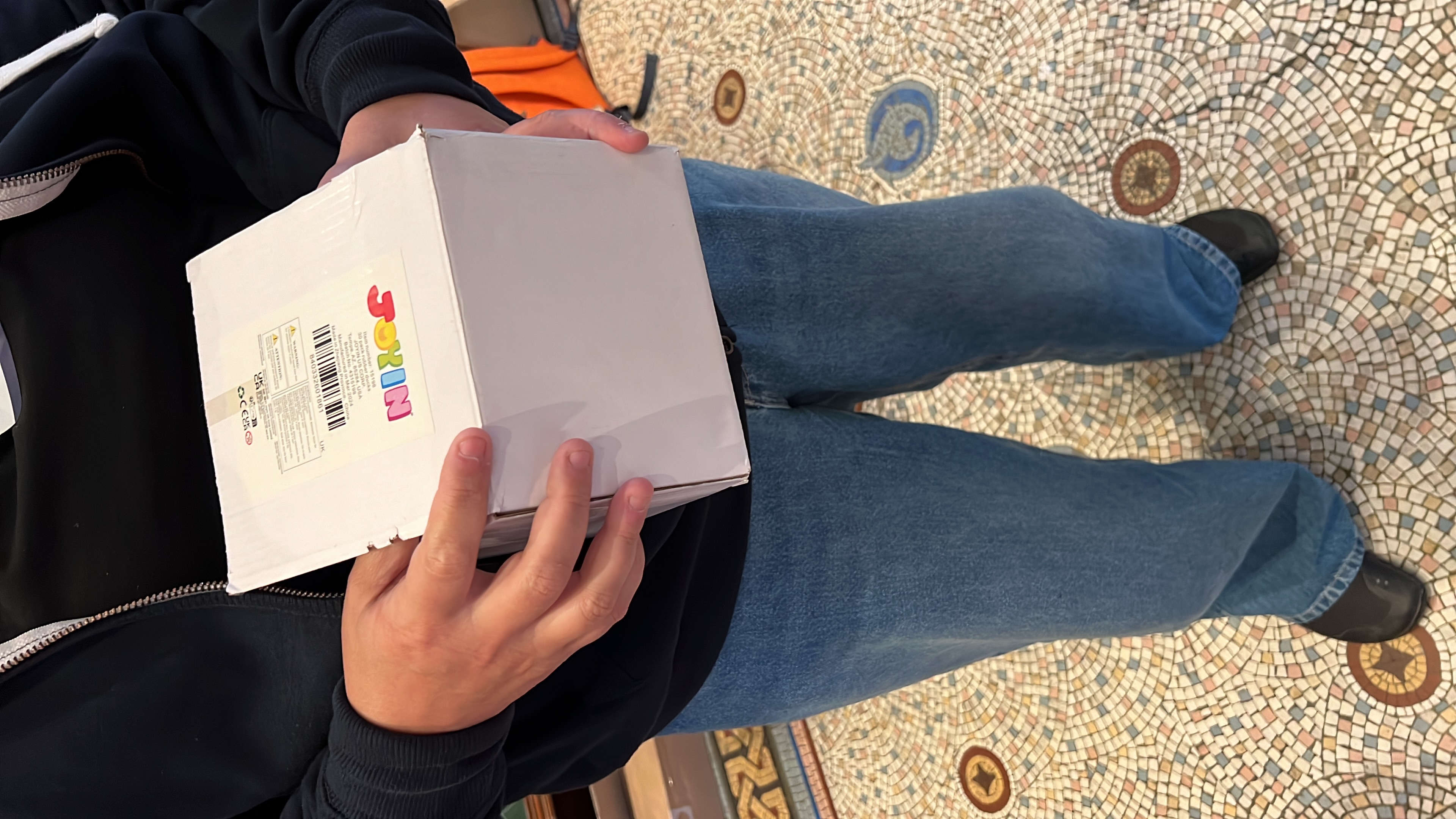 The bottom two-thirds of a person holding a white box full of small toy ducks. 