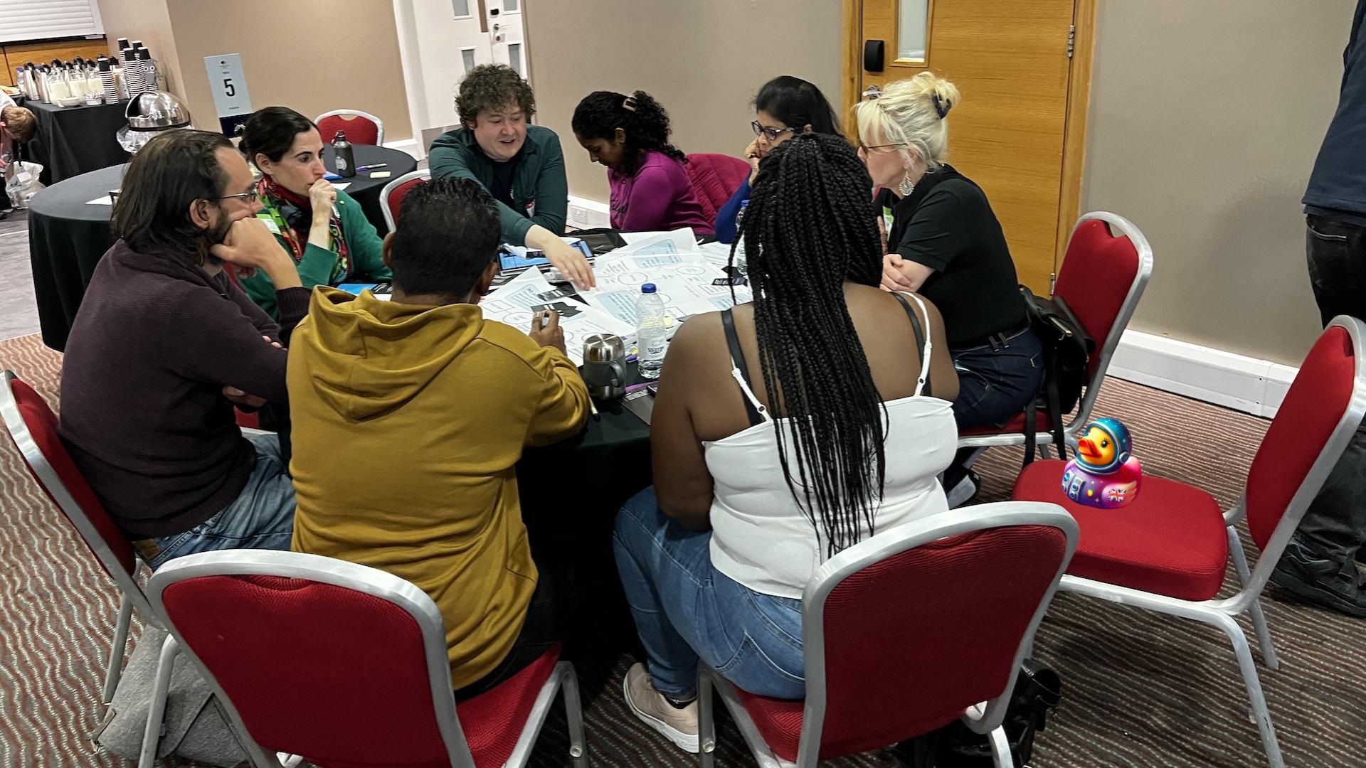 A group of eight people sat around a circular table. They are in deep discussion running through a security testing exercise. A bug and duck character have been added to the photo. Bug is harder to find than duck.