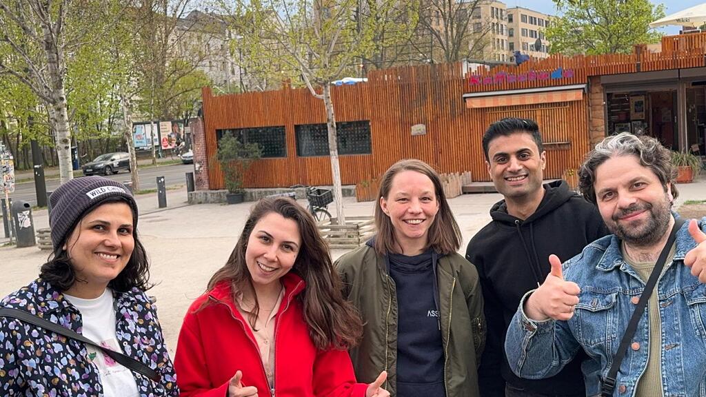 Five people standing outdoors in a park-like urban area, smiling at the camera. Two people in the front are giving th... image