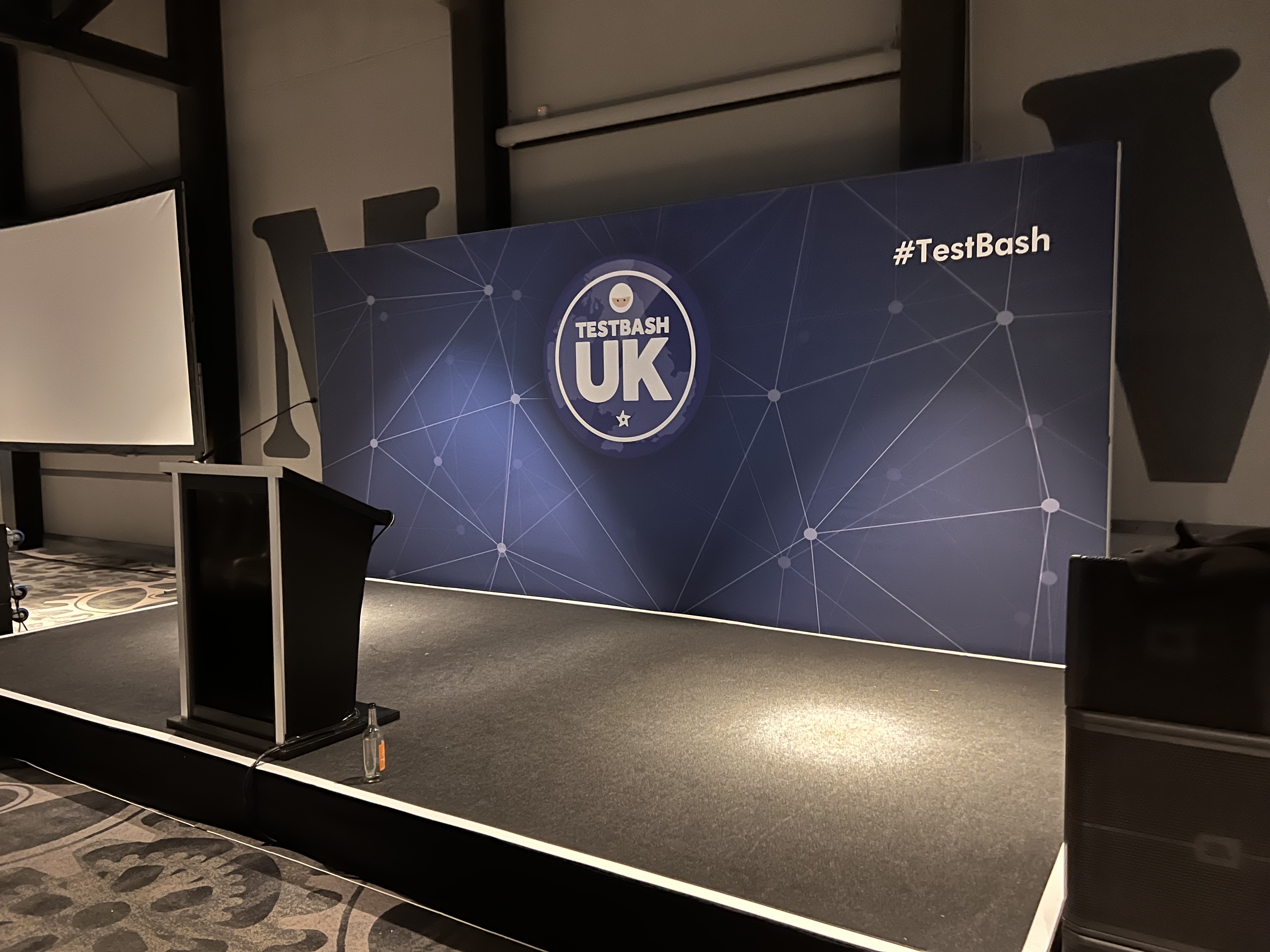 An empty event stage with black carpeting, featuring a dark podium on the left and a large blue backdrop board. The b... image