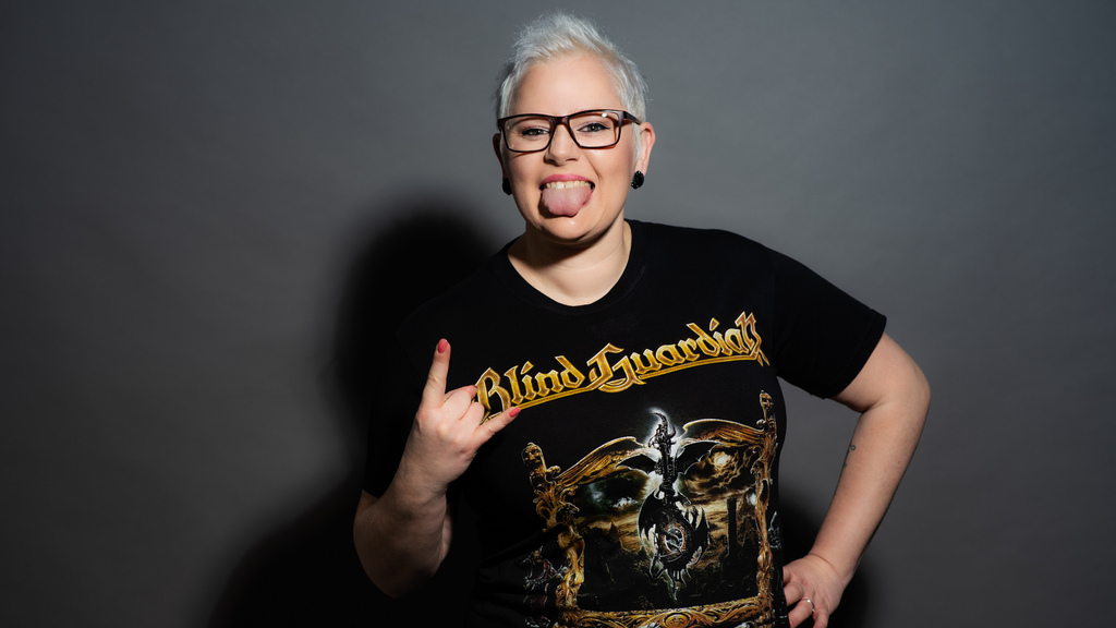 Studio photo of a woman with short platinum hair and glasses, playfully sticking out her tongue and flashing the rock hand sign, wearing a black “Blind Guardian” band T-shirt against a gray backdrop