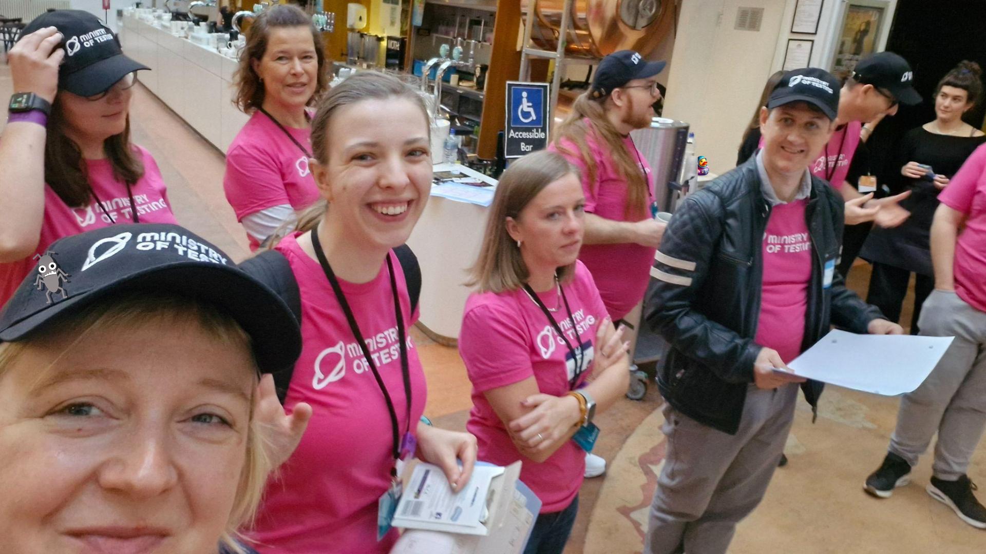 A selfie of a group of eight or so people. They have matching pink t-shirts with Ministry of Testing logos on. Some are wearing MoT branded black caps. A small bug and small duck character have been added to the photo. 