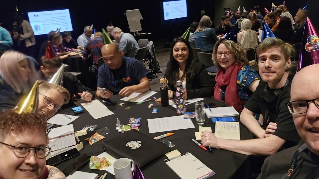 Group of people all wearing party hats, sitting round a table at a MoTaCon workshop. image