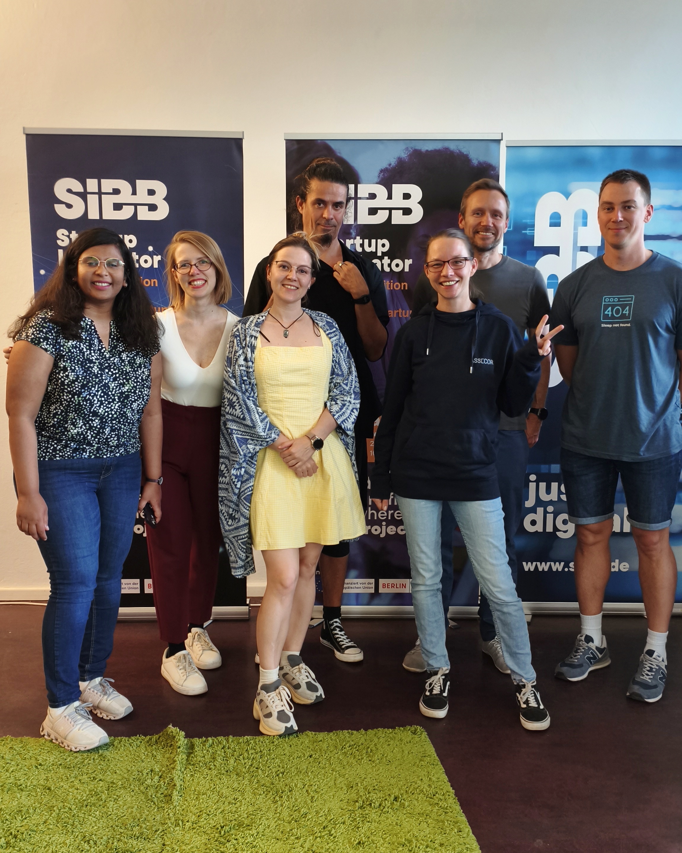 A group of seven people smiling and standing in front of two SIBB Startup Incubator banners. They are posing casually indoors on a dark floor with a small green rug at their feet. The group includes men and women, dressed casually in jeans, sneakers, dresses, and hoodies, showing a friendly and relaxed vibe from a community event.