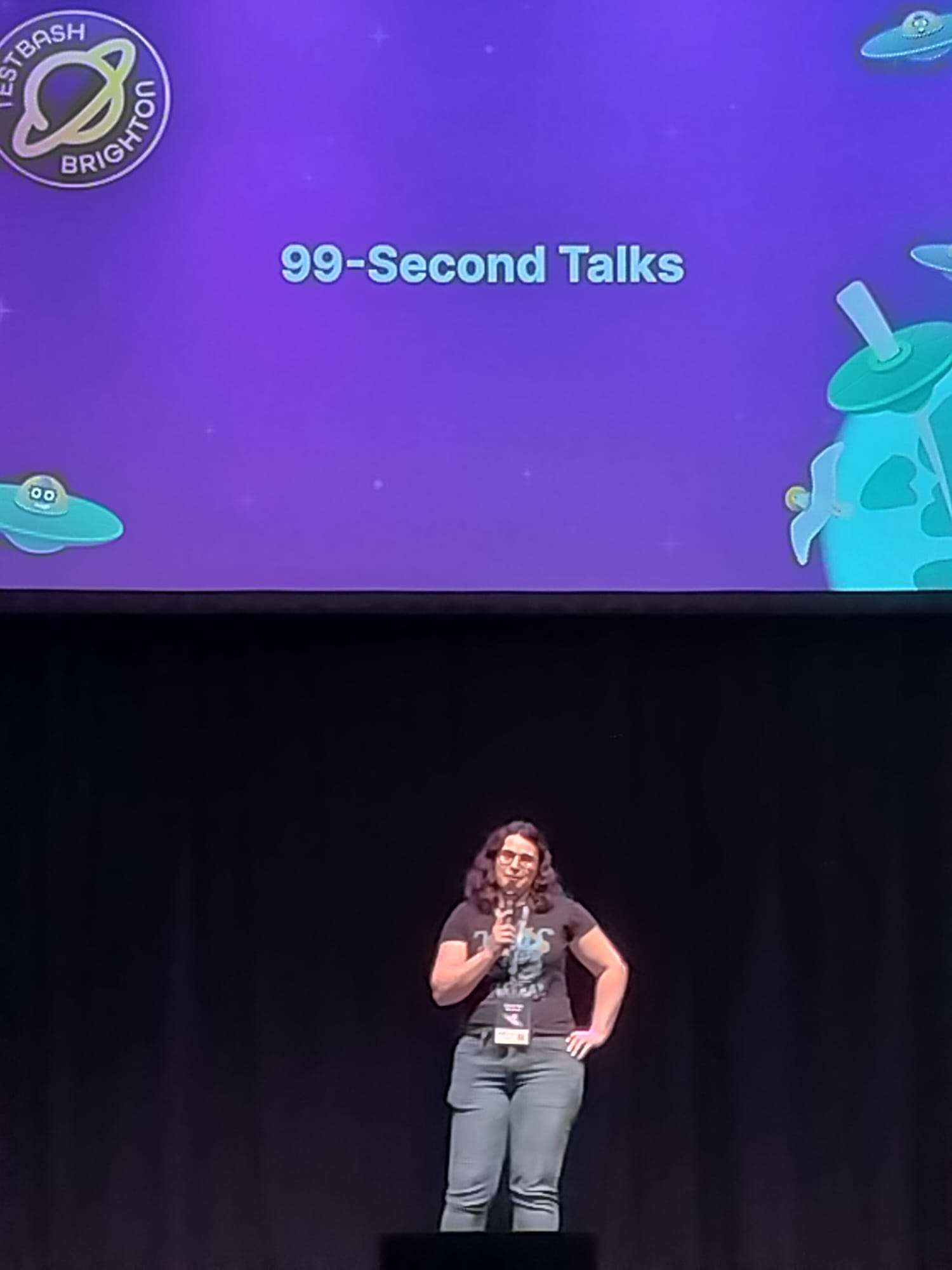 A person wearing jeans and a black t-shirt on stage at MotaCon 25, giving a 99-second talk.