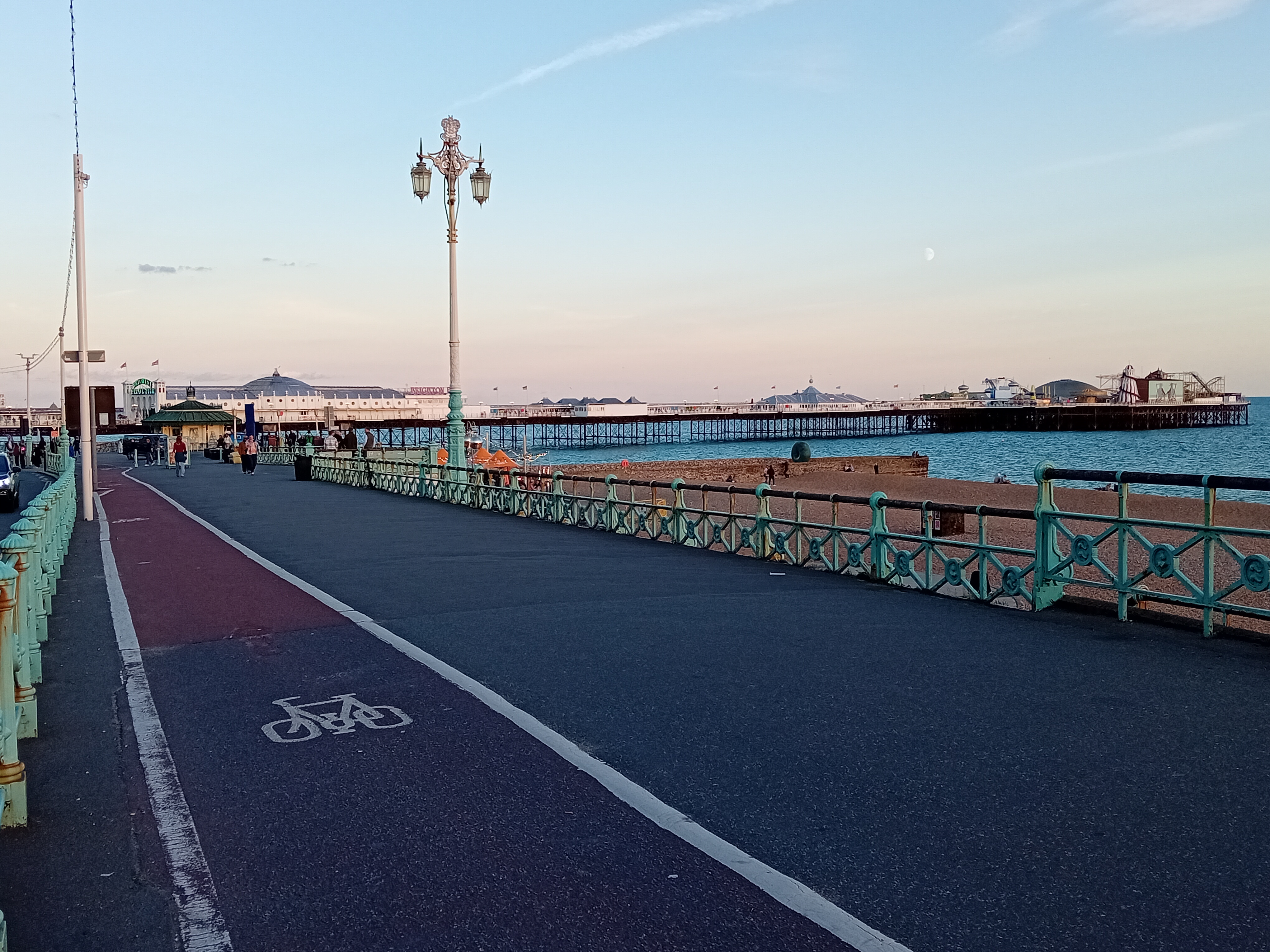 A photograph of the Promenade of Brighton Seafront.  In the picture there is a pavement with a cycle lane.  To the right of the pavement are green railings and a lamp post and then a beach and the sea.  At the back of the photograph is a pier and to the right there is a beach and the sea.  It is dusk and the sky in the picture is clear.
