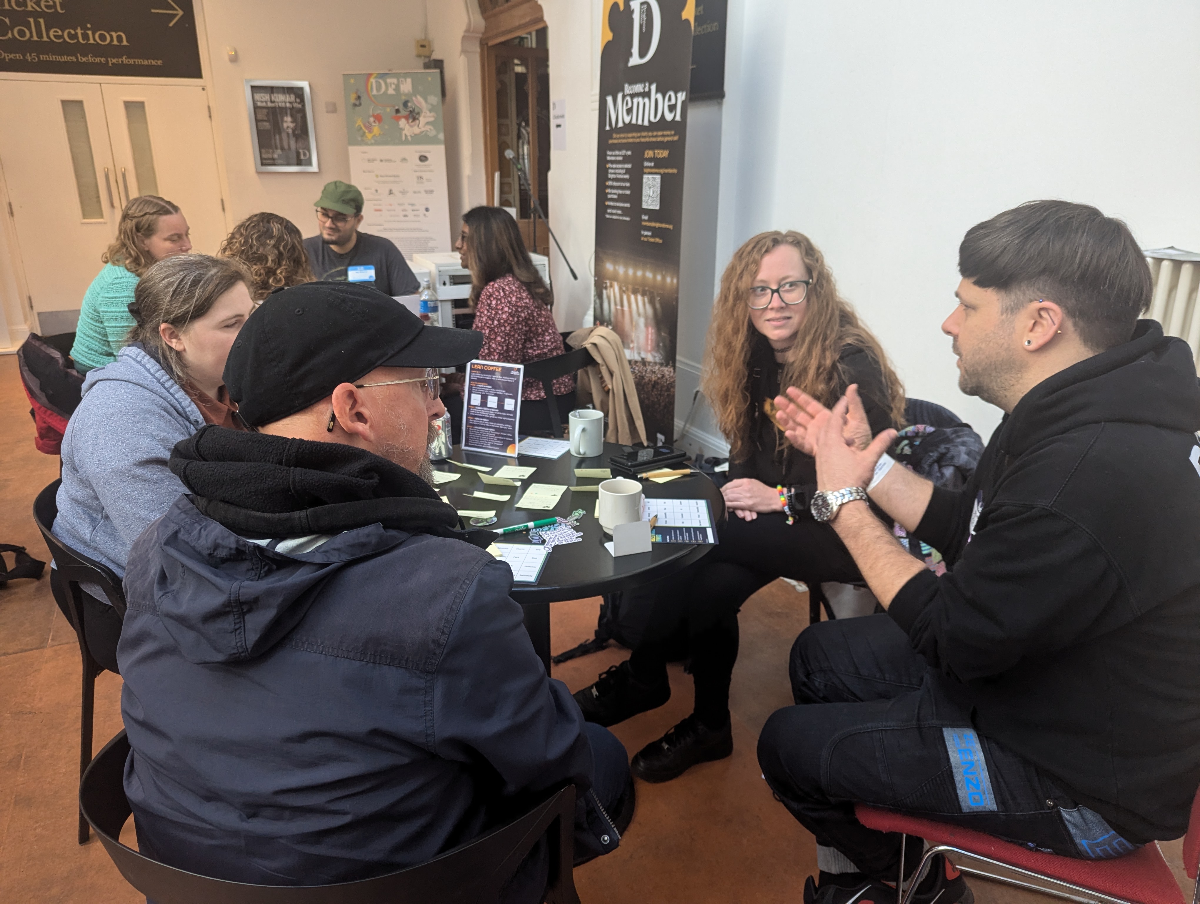 A group of people participating in a Lean Coffee session at TestBash 2024. They are all seated around small round tables, engaged in discussion and exchanging ideas related to software testing. Sticky notes and materials for organizing the session are spread across the table. 