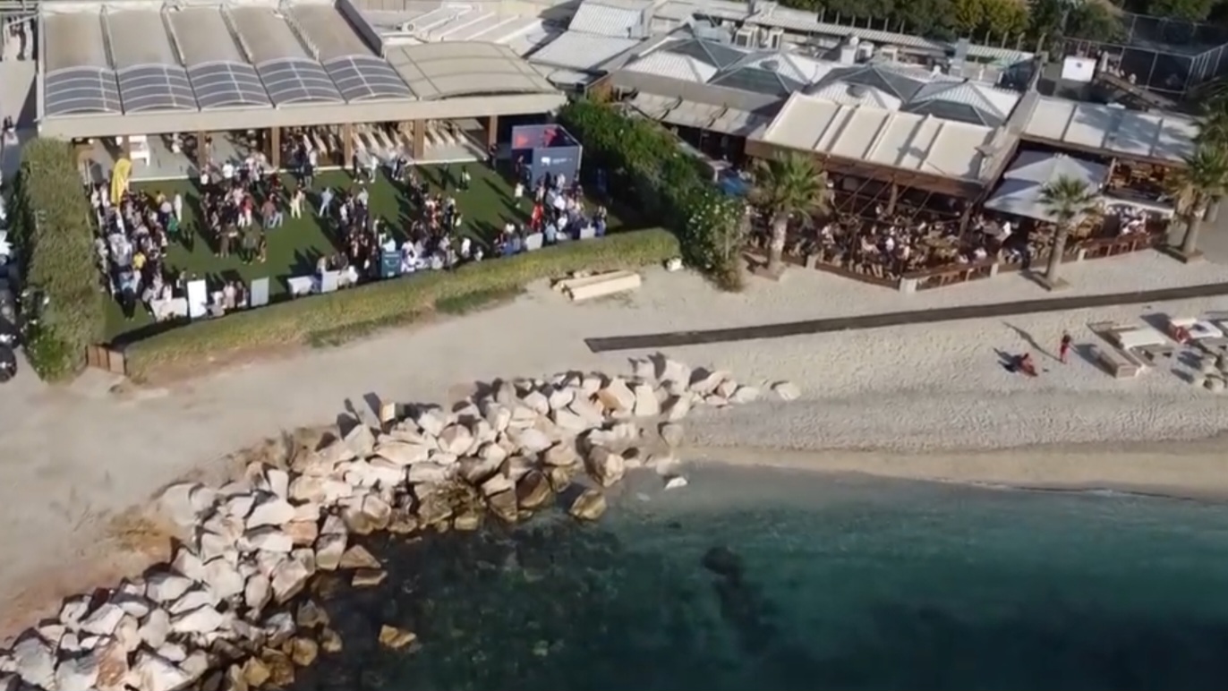 An aerial shot of the Ministry of Testing Athens venue with numerous people stood outside in conversations. The venue is very bright (sunny) and is located directly infront of the beach.
