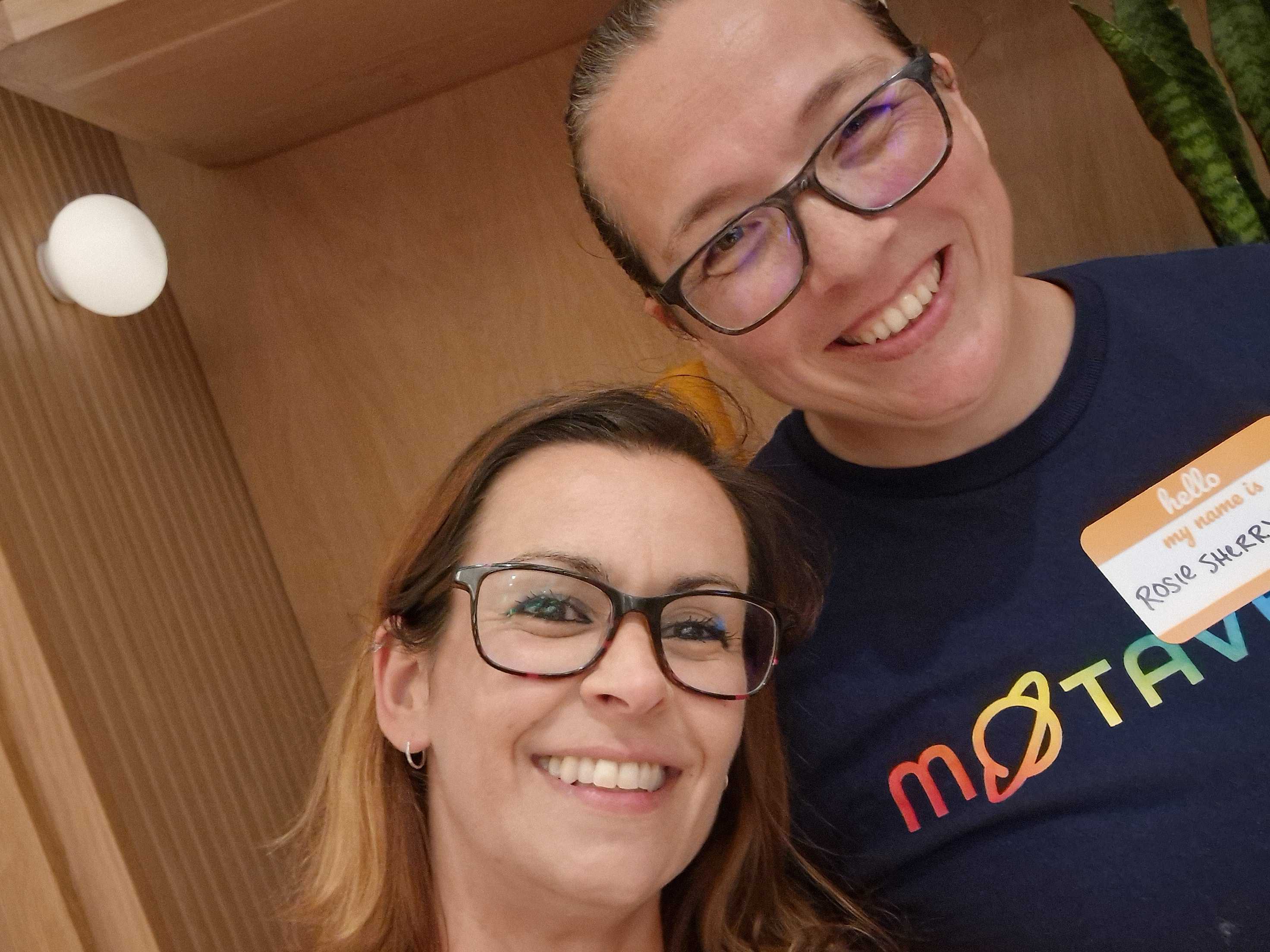 Close-up selfie of two smiling women wearing glasses. The person on the right wears a navy blue t-shirt featuring the... image