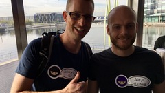 A selfie of two white men in an event space. There is a water in the background.  image