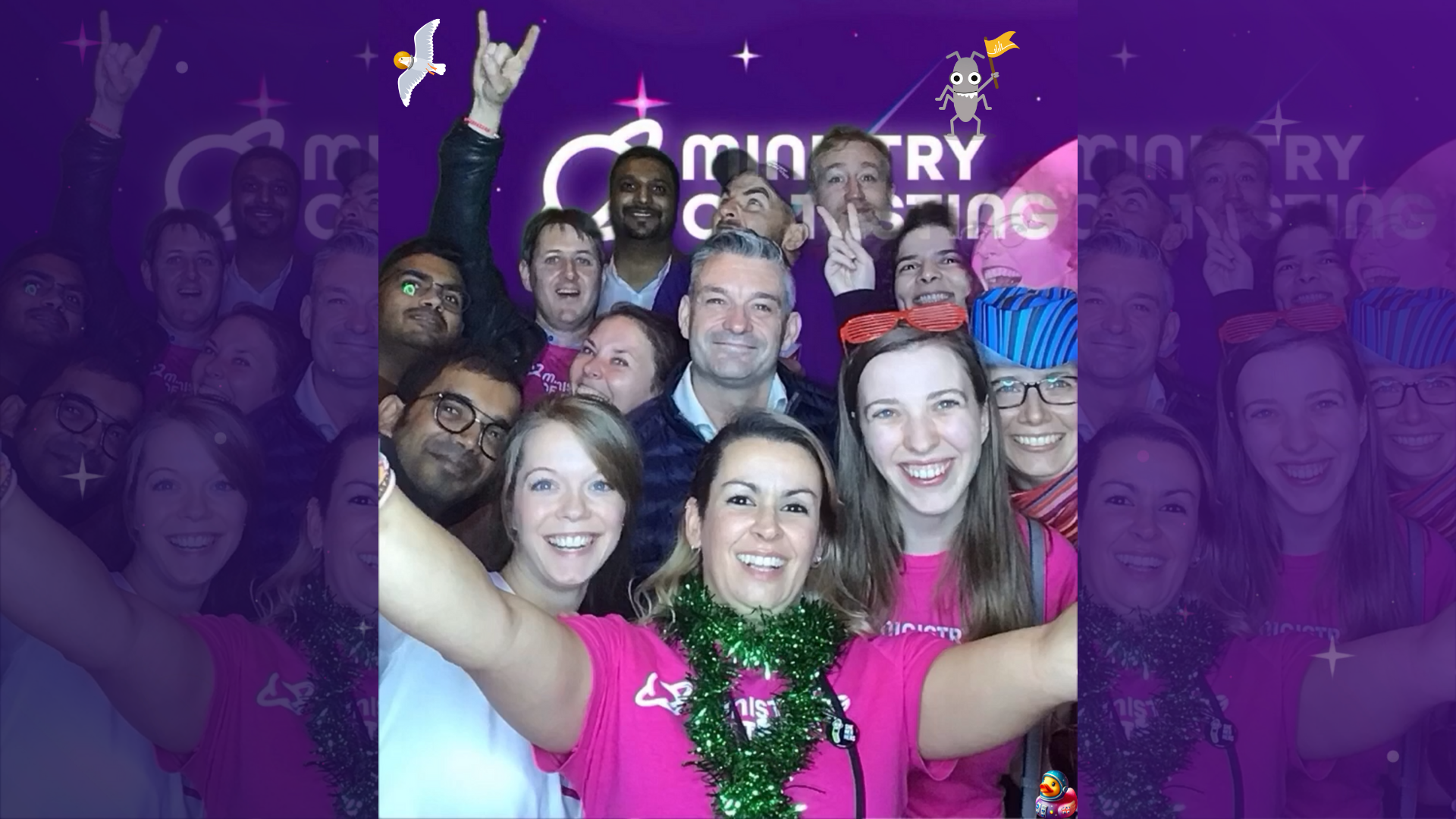 A large group of adults are smiling and posing for a selfie in a photobooth in front of a backdrop that says "Ministry of Testing". The people in the foreground are wearing pink t-shirts, one with a green tinsel garland around her neck. Some individuals in the background are making hand gestures. The lighting appears to be from a flash, creating some highlights on faces. The overall atmosphere seems joyful and celebratory.