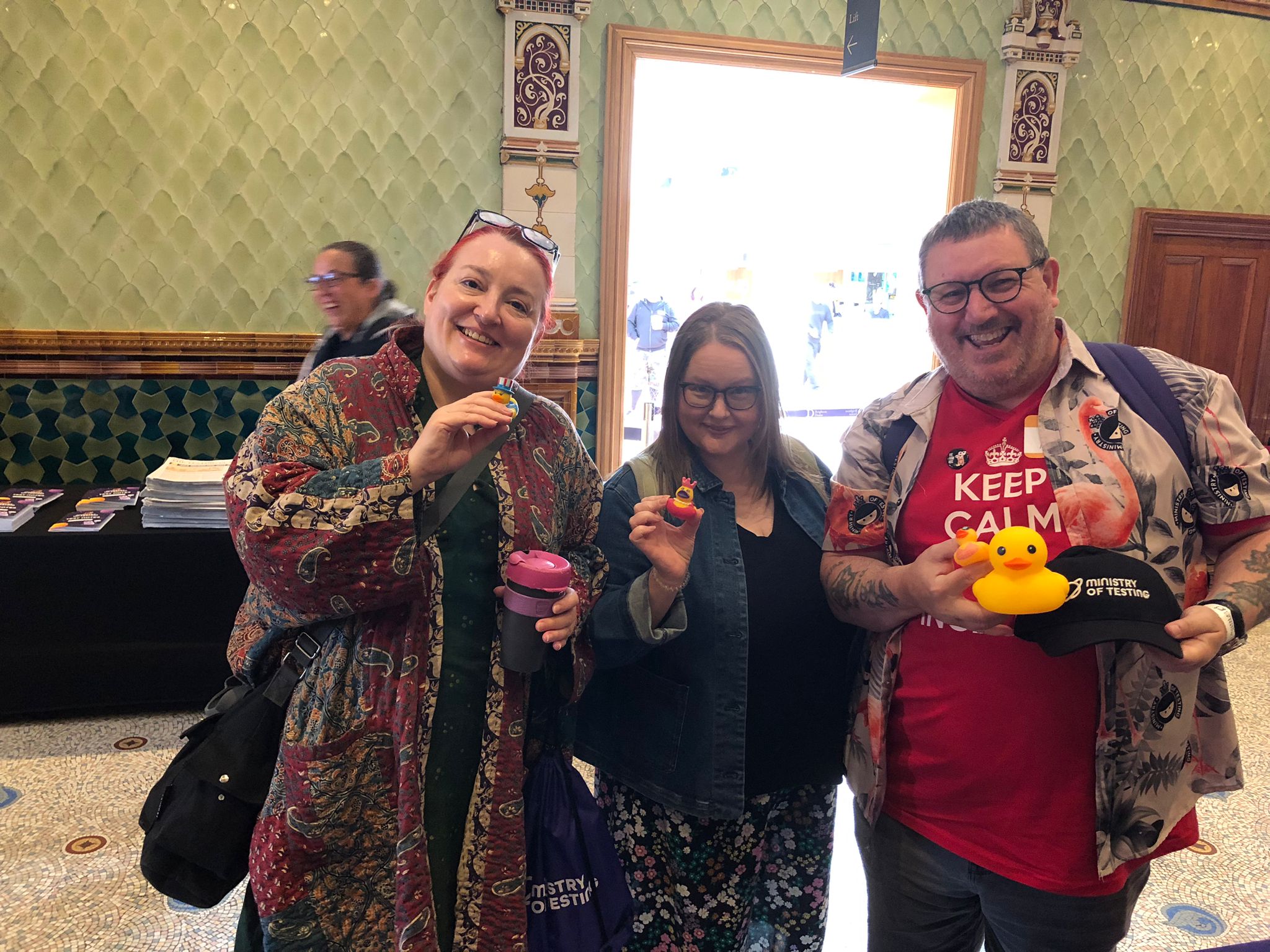 A picture with three TestBash attendees showing the ducks they found around the venue and some of the swag they swapped it for. One of the three attendees is Ady Stokes who is wearing a 'Keep Calm' red t-shirt. In the background, we can see Rosie Sherry passing behind them and laughing whilst out-of-focus.