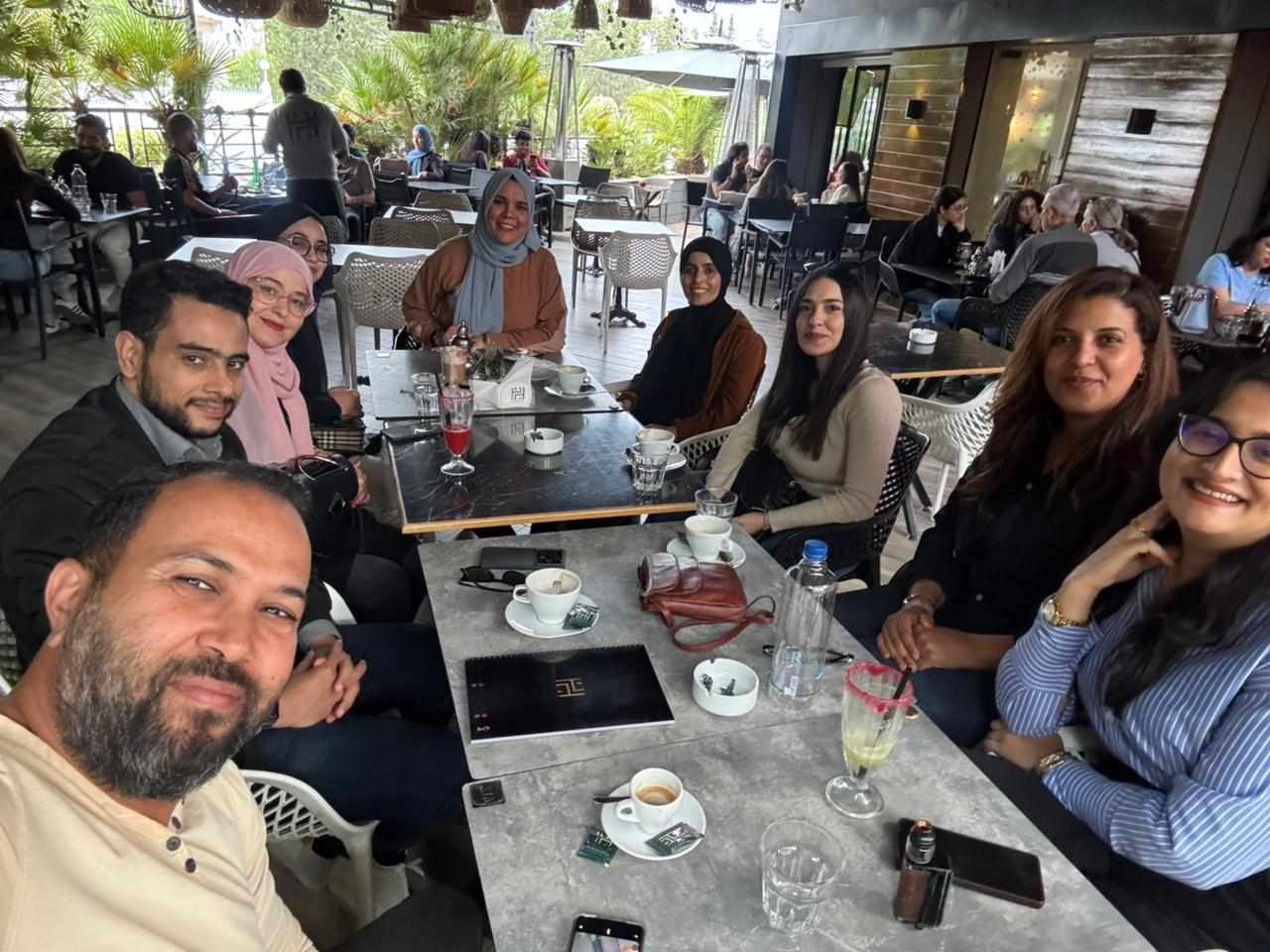 A group of nine people, including both men and women, are gathered around a table at an outdoor café. They are smiling and appear to be enjoying a casual meet-up or discussion. On the table are coffee cups, glasses, notebooks, a water bottle, and a purse. The background shows other patrons seated at tables, lush greenery, and a shaded terrace area with natural light filtering in.