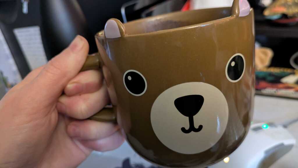 Left hand badly holding a mug of coffee. The mug is a light brown chunky mug with black eyes and a cute mouth with a ... image