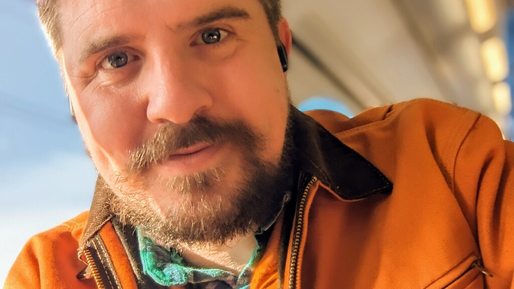 Head and shoulder picture of Scott on a train , wearing a brown jacket, With sunset filter and pink and blue hair fresh for test bash 