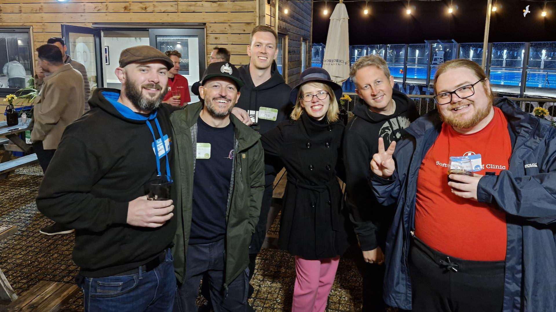 A group of six people smiling at the camera. They are outside socialising in an event space. There is an outdoor swimming pool in the background. A bug, duck and seagull character are attempting to hide in the photo.