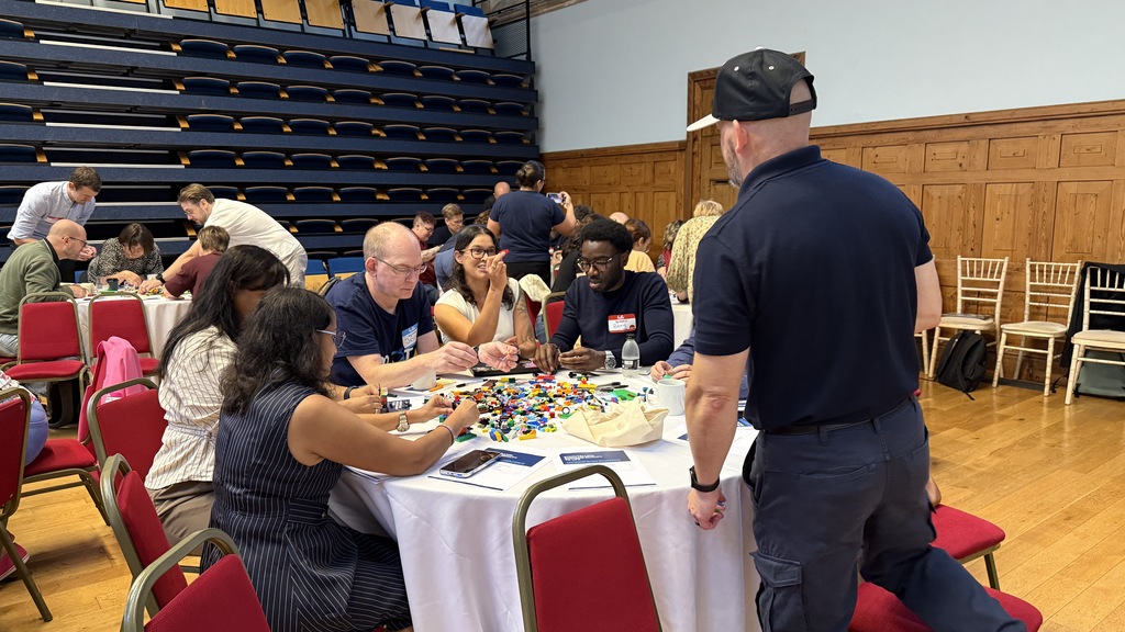 A room of quality professionals on three tables. In the foreground a group at a table are building with Lego. The Leg... image