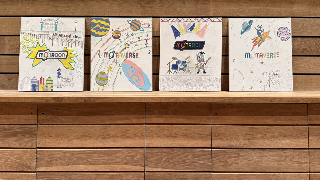 Four colorful hand-drawn art canvases featuring "MOTACON" and "MOTAVERSE" logos sit lined up on a wooden shelf agains... image