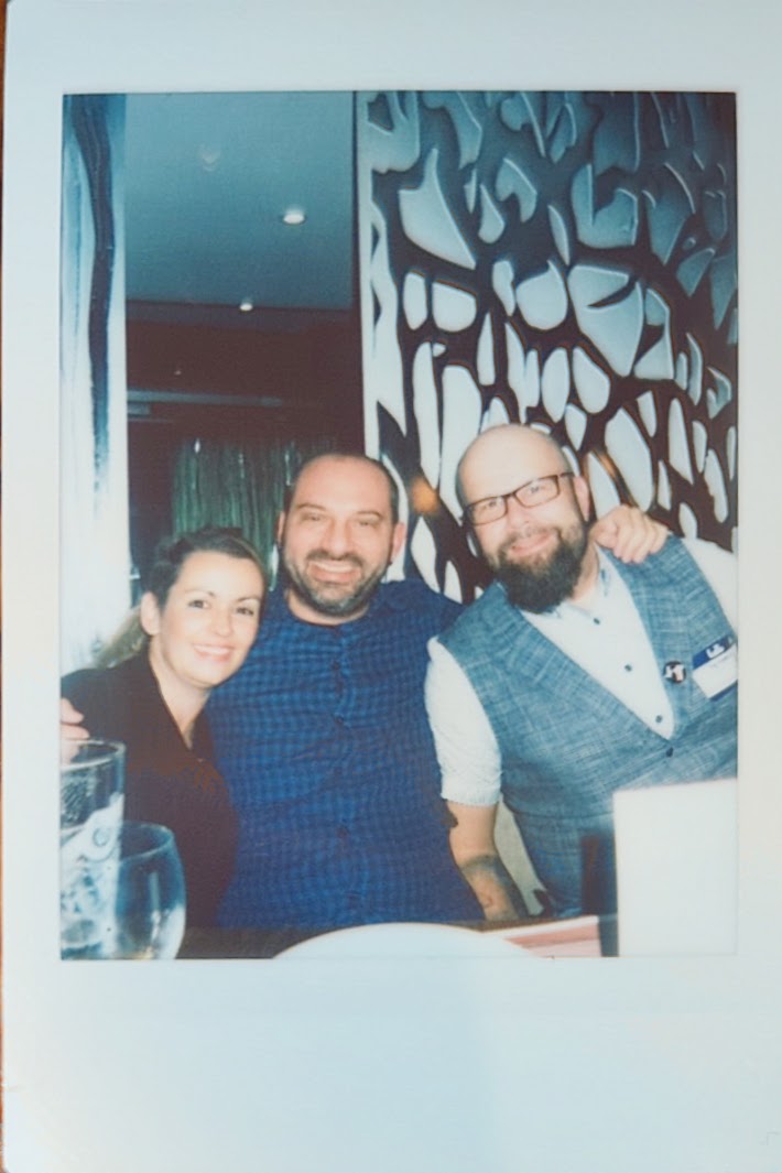 Diana, Petros, and Christian smiling together at a dinner table, with a decorative wall in the background.