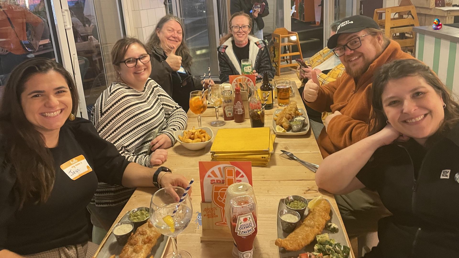 A group of seven adults smiles around a wooden table laden with fish and chips and drinks in a brightly lit restaurant. A seagull, duck and bug character have been added to the image. 