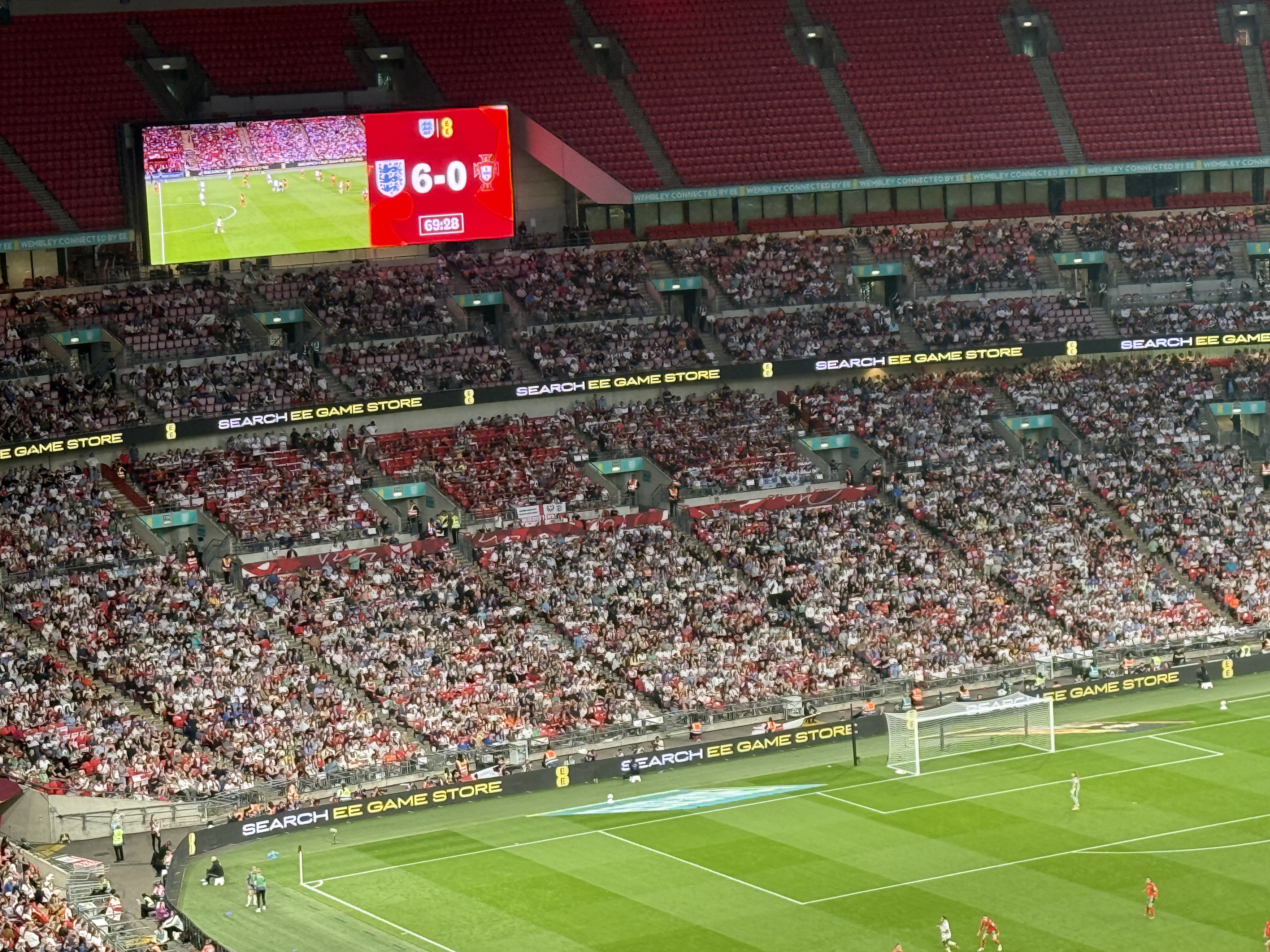 The crowd at Wembley stadium. There's a football pitch with a goal. The score is 6 to England and 0 to Portugal. 