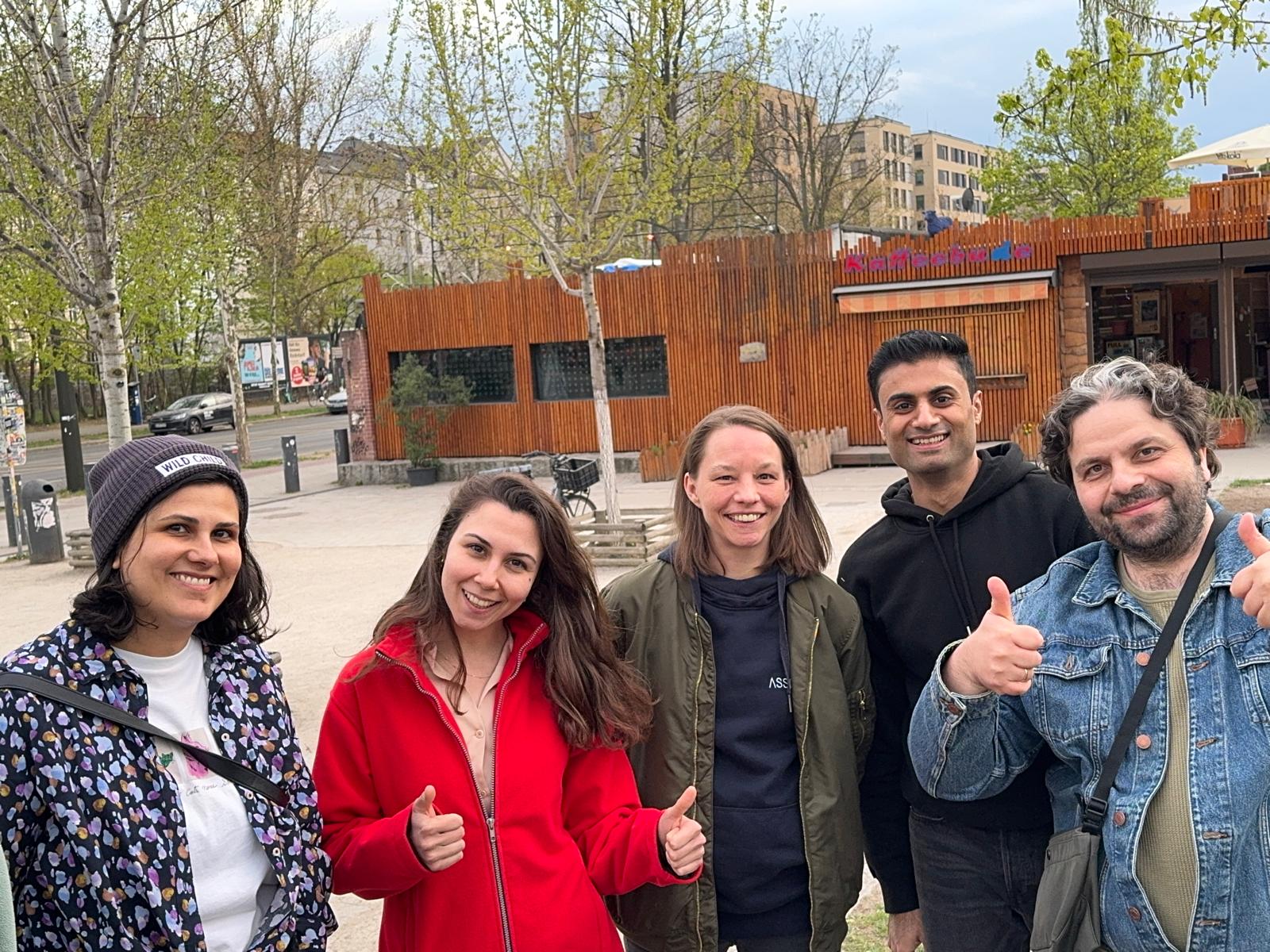 Five people standing outdoors in a park-like urban area, smiling at the camera. Two people in the front are giving th... image