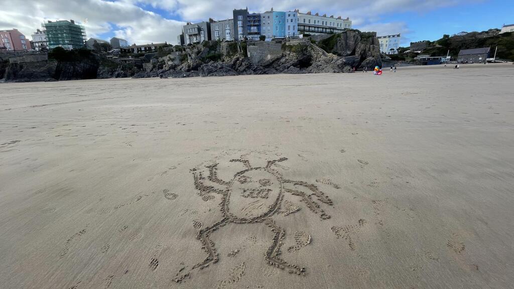 A cartoonish drawing of a bug or alien creature with six legs and antennae, etched into the sand on a beach. In the background, beyond the wide expanse of sand, a rocky outcrop is topped with a row of colourful, multi-story buildings under a partially cloudy sky. A bug, a space seagull and a space duck character have been added to the photo. 