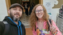 A bearded man in a cap and a woman with glasses and long red hair take a selfie indoors. The woman wears a pink jacket and a name tag while flashing a peace sign. People are visible in the backgrou... image