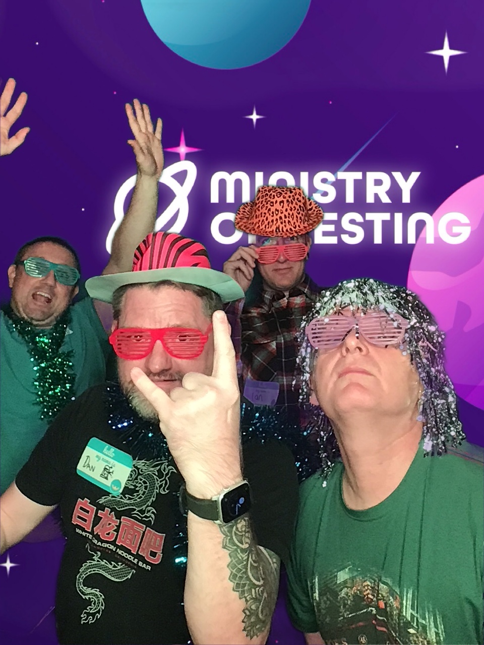 This pic depicts a 4 person TestBash Photo Booth moment. Attendee no1 is at the back left and has both hands in the air and look a bit like they are on a rollercoaster, they are also wearing all green including the decorative accessories from the dressing up table. Attendee no2 is closer to the camera and more in the centre they are throwing up a 'rock on' sign and you can just about make up a name tag on their right chest that says DAN. Attendee no2 has on red accessories in the form of plastic cut out glasses and a red zebra print hat. Attendee no3 is a little more towards the back and is peering at us over their orange cut out glasses whilst wearing an orange leopard print hat and a checkered plaid shirt. Attendee no4 is right at the front and to the right and wears a silver tinsel wig, pink cut out glasses and is peering at us through the cut-out glasses,. He is also wearing green. 

Background: The green screen backdrop depicts a simple illustrated outer space scene. The main colour is purple, with cute stars and planets in shades of pink, blue, and white. About a third of the way down the screen, a logo appears. It reads "Ministry of Testing" in bold white text with a blue shooting star behind it. Next to the text, in the same bold white style, is a line drawing of the planet Saturn.