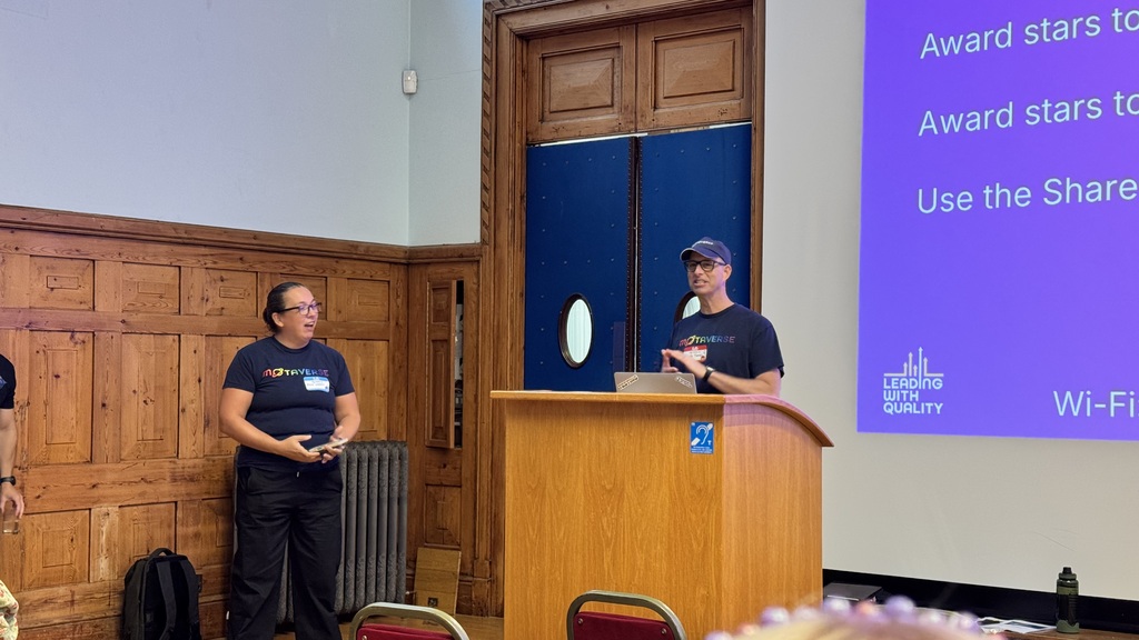 Rosie stands smiling beside a wooden lectern while Simon, wearing glasses and a baseball cap, speaks to the room. Bot... image