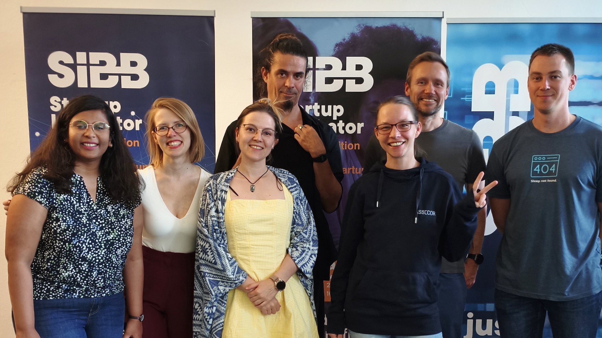 A group of seven people smiling and standing in front of two SIBB Startup Incubator banners. They are posing casually indoors on a dark floor with a small green rug at their feet. The group includes men and women, dressed casually in jeans, sneakers, dresses, and hoodies, showing a friendly and relaxed vibe from a community event.