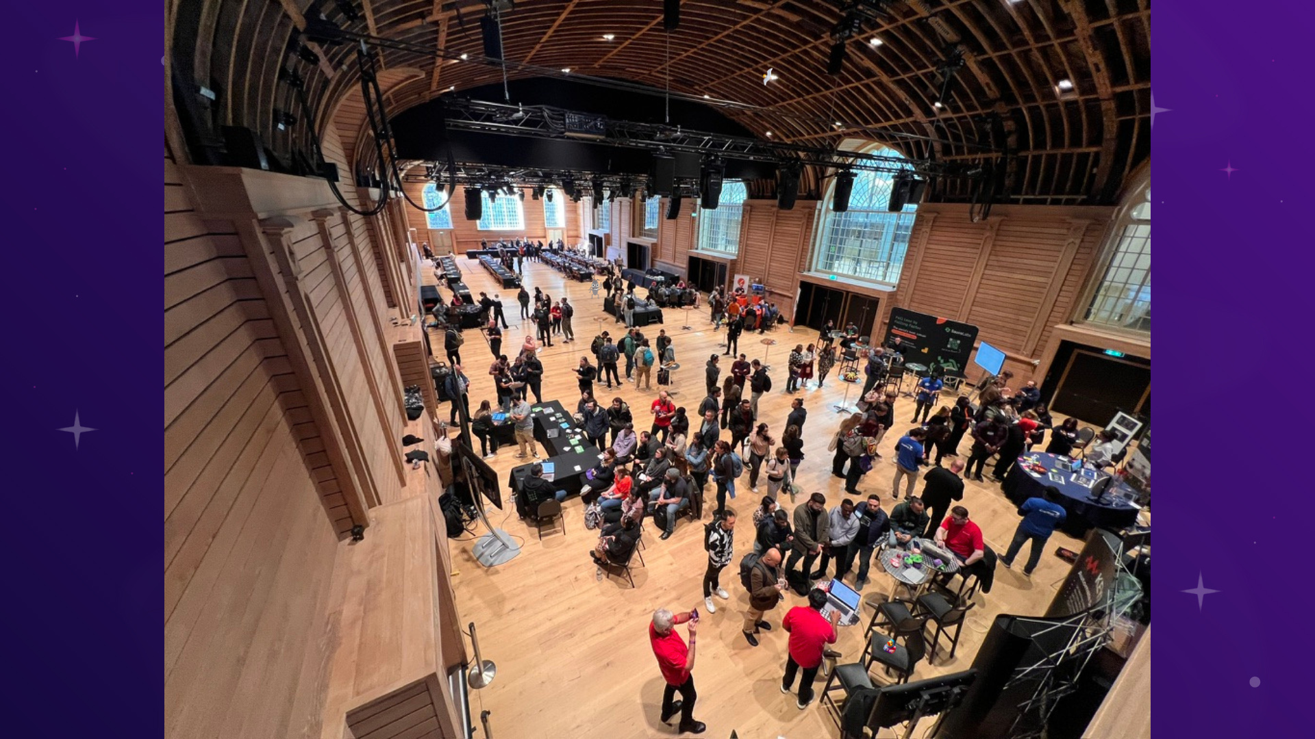 A high-angle view of a large, high-ceilinged hall filled with people attending a tech event. The hall has wooden panelling and large windows, with several tables and booths set up. People are standing around, talking, and interacting with the exhibits. A bug, a seagull and a duck character have been added to the photo. 