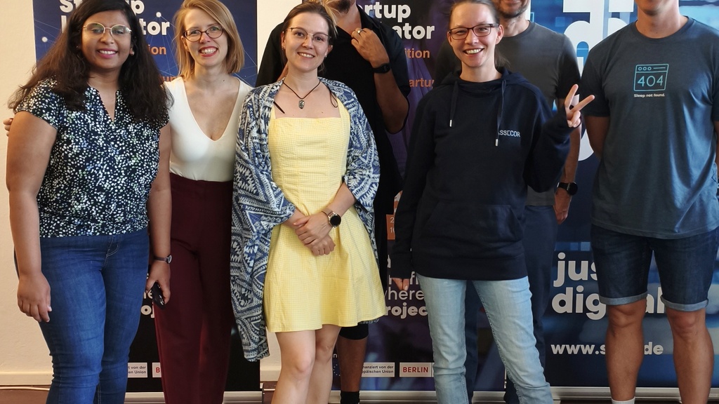 A group of seven people smiling and standing in front of two SIBB Startup Incubator banners. They are posing casually indoors on a dark floor with a small green rug at their feet. The group includes men and women, dressed casually in jeans, sneakers, dresses, and hoodies, showing a friendly and relaxed vibe from a community event.