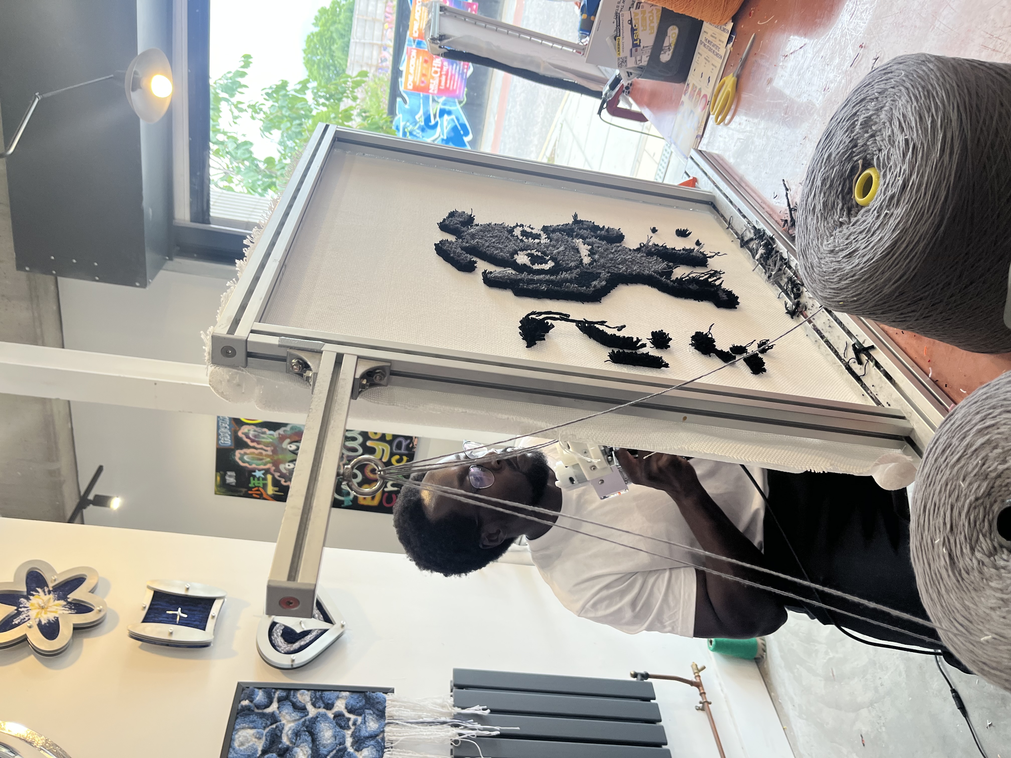 A picture of Eamon Droko at Cheeky studio who is rug tufting a rug of Bug using the classic grey, black and white colours. He is focused and concentrating whilst using the rug tufting gun. 