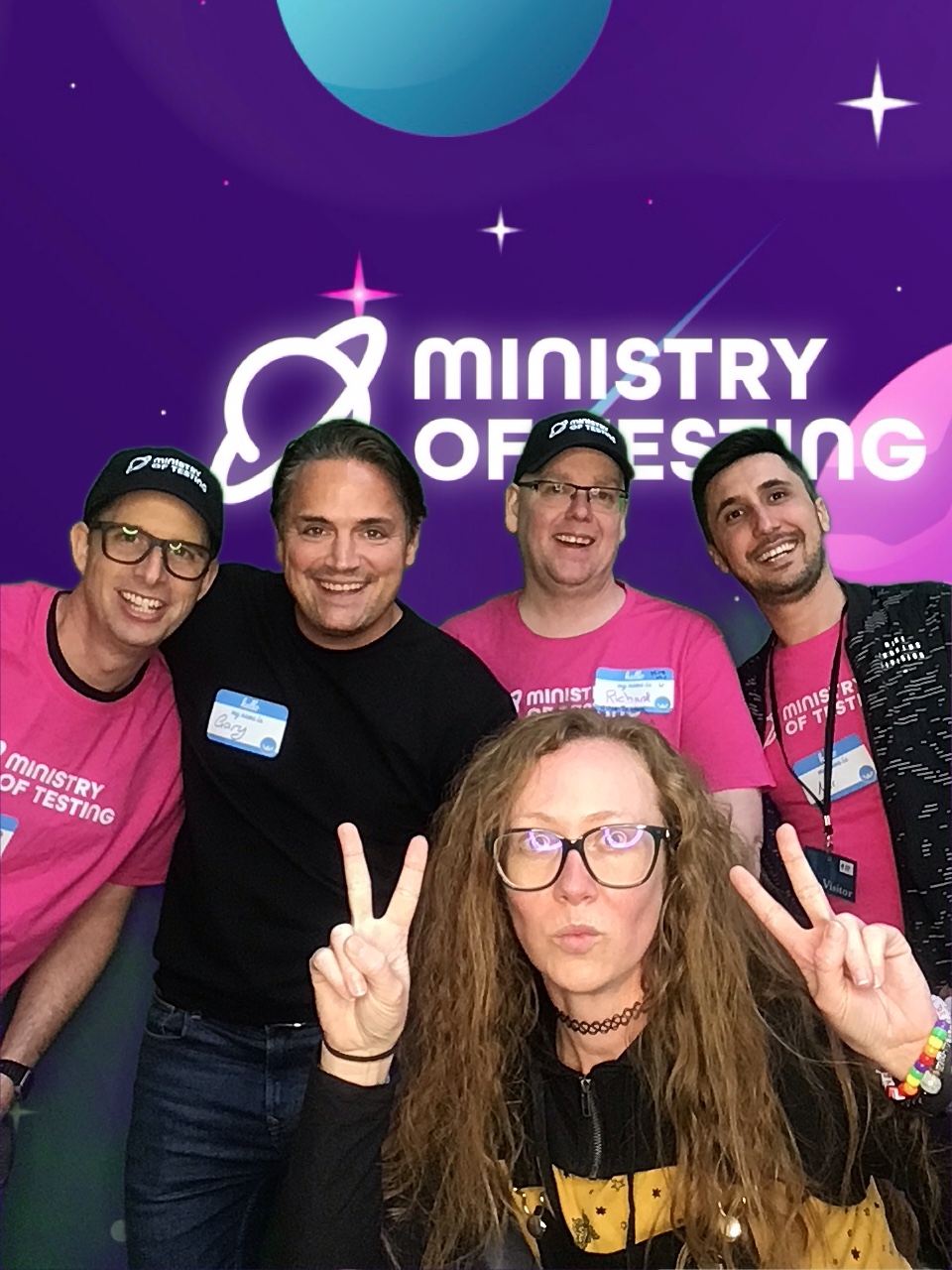 5 attendees rush the photobooth for a high energy photo.

No silly accessories in this picture but plenty of Ministry of Testing apparel can be seen. 3/5 attendees have pink MoT tees on and the others are wearing black, 2 of the attendees also have black MoT baseball caps on. 

Smiles all round! 

Background: The green screen backdrop depicts a simple illustrated outer space scene. The main colour is purple, with cute stars and planets in shades of pink, blue, and white. About a third of the way down the screen, a logo appears. It reads "Ministry of Testing" in bold white text with a blue shooting star behind it. Next to the text, in the same bold white style, is a line drawing of the planet Saturn.